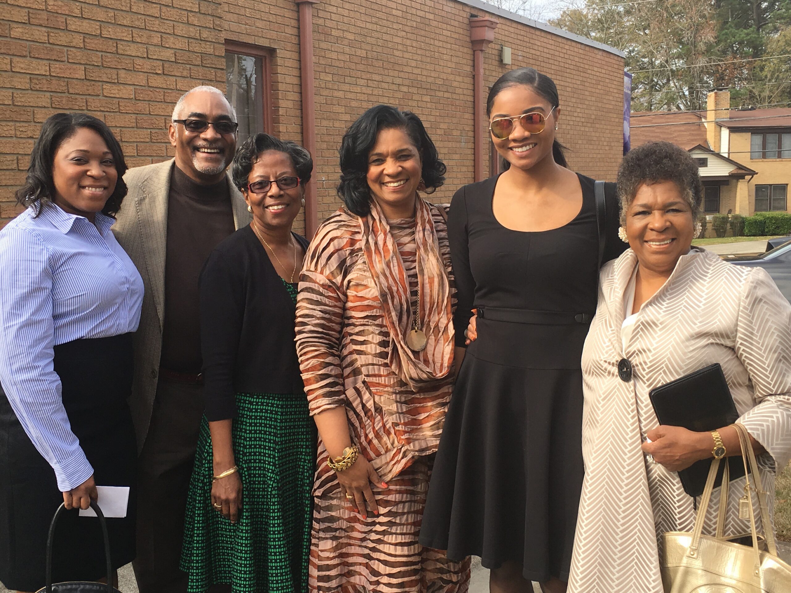 Group of six adults posing together outside a brick building, smiling at the camera; sunny day, casual/formal attire.