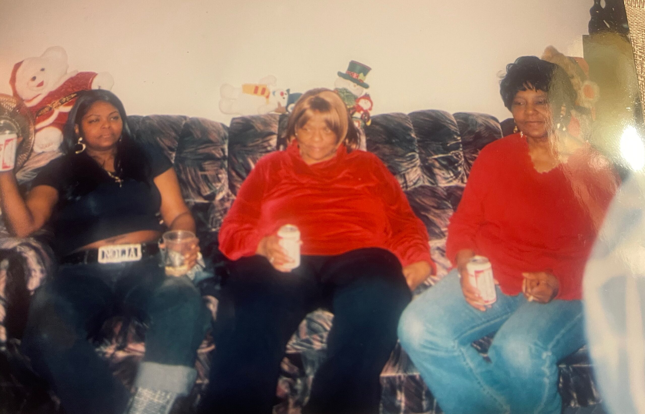 Three women seated on a dark patterned sofa with Christmas decorations in the background, each holding a cup or drink.
