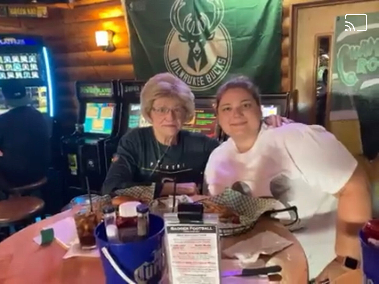 Two women sit at a neon-lit bar with drinks, smiling at the camera; older woman with glasses on the left and a younger woman in a white shirt on the right, arcade machines in the background.