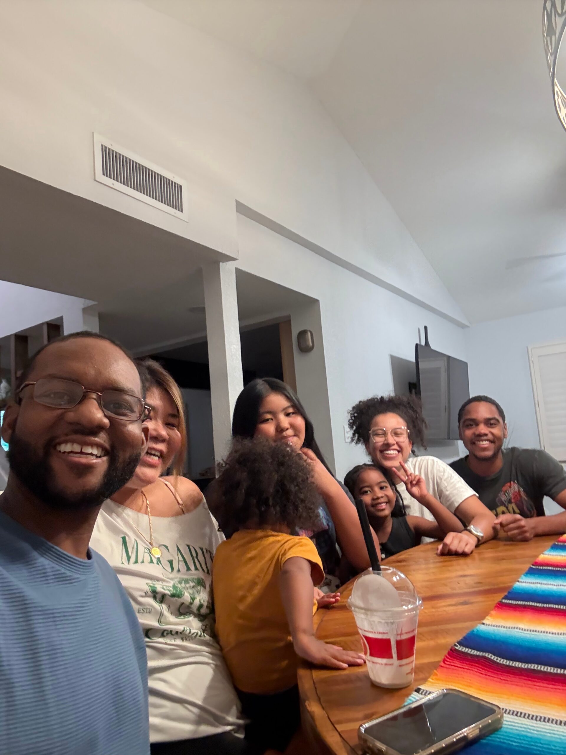 Group of friends around a wooden table, smiling for a selfie in a bright room with beverages and a colorful striped table runner.
