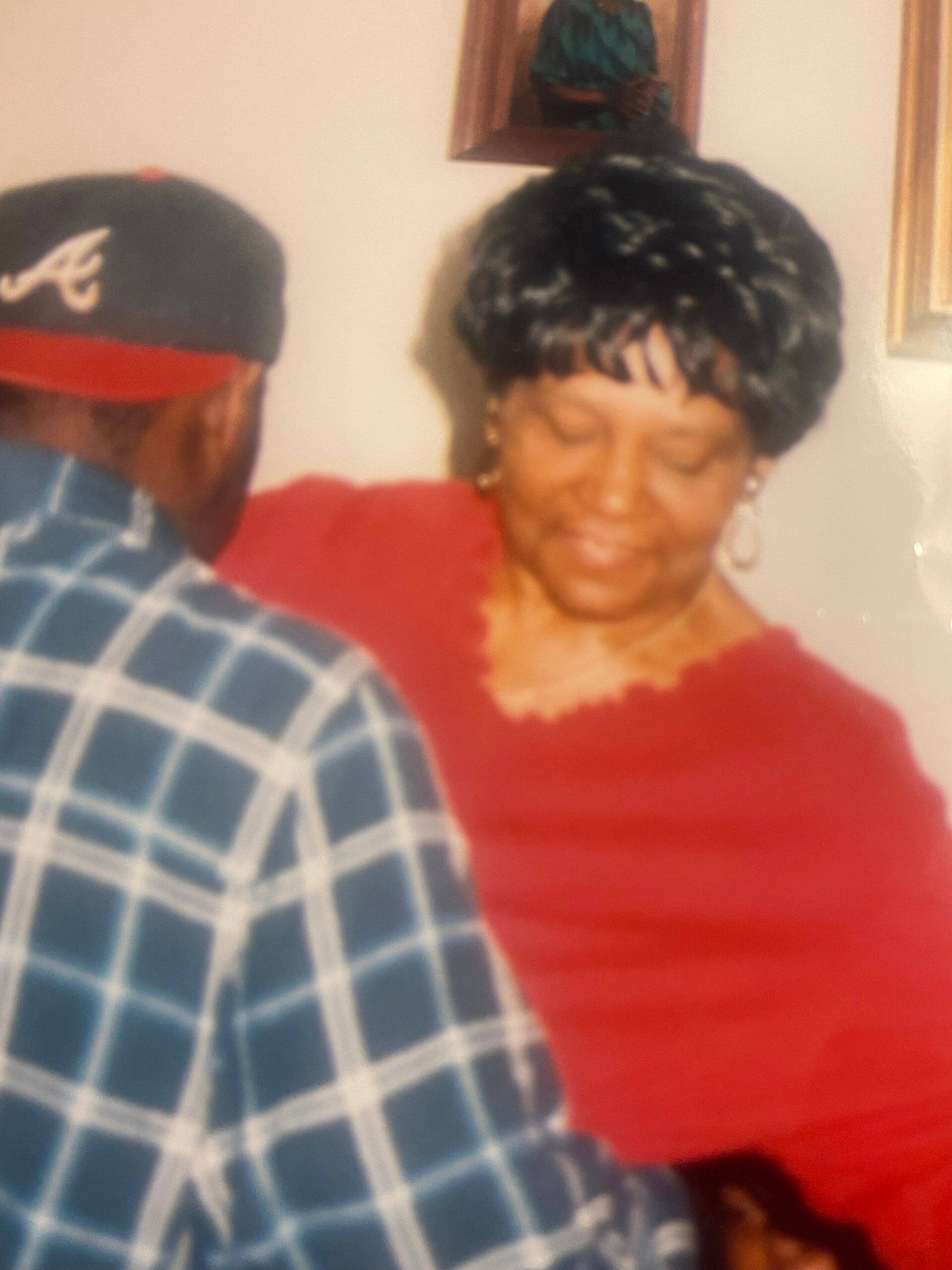 Blurred photo of an elderly woman in a red top smiling, sitting with a man in a blue plaid shirt indoors.