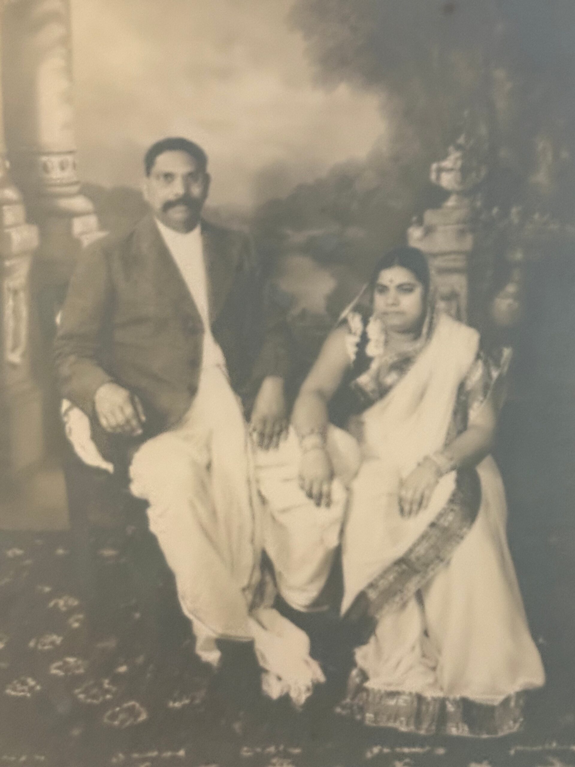 Vintage photo of a man in a suit and a woman in a traditional sari with garlands, posing together outdoors on a cloudy day.