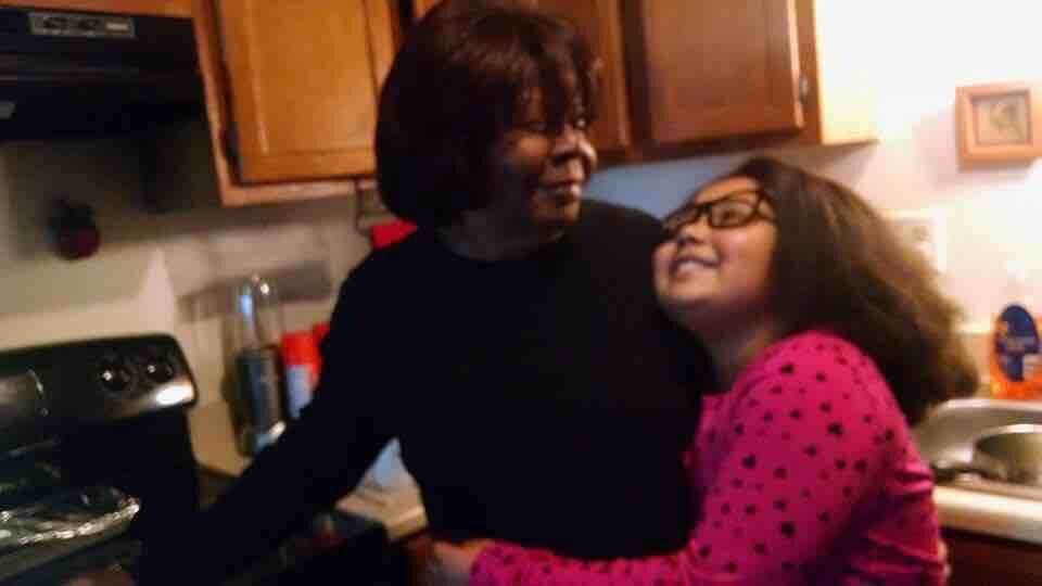 Two people embrace and smile in a home kitchen, one in a black top and the other in a pink polka-dot sweater.
