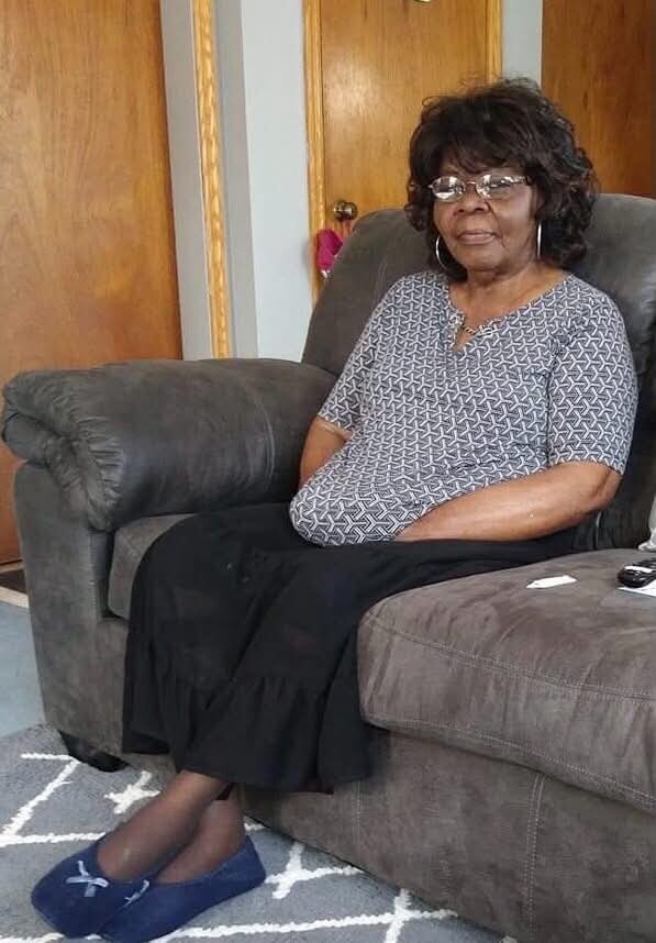 Older woman wearing a patterned blouse and black skirt sits on a gray sofa with her hands in her lap, in a living room setting.