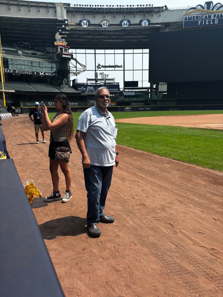 Older man wearing a light gray polo and jeans stands on the infield dirt at American Family Field, while a young woman takes his photo behind him in the background.