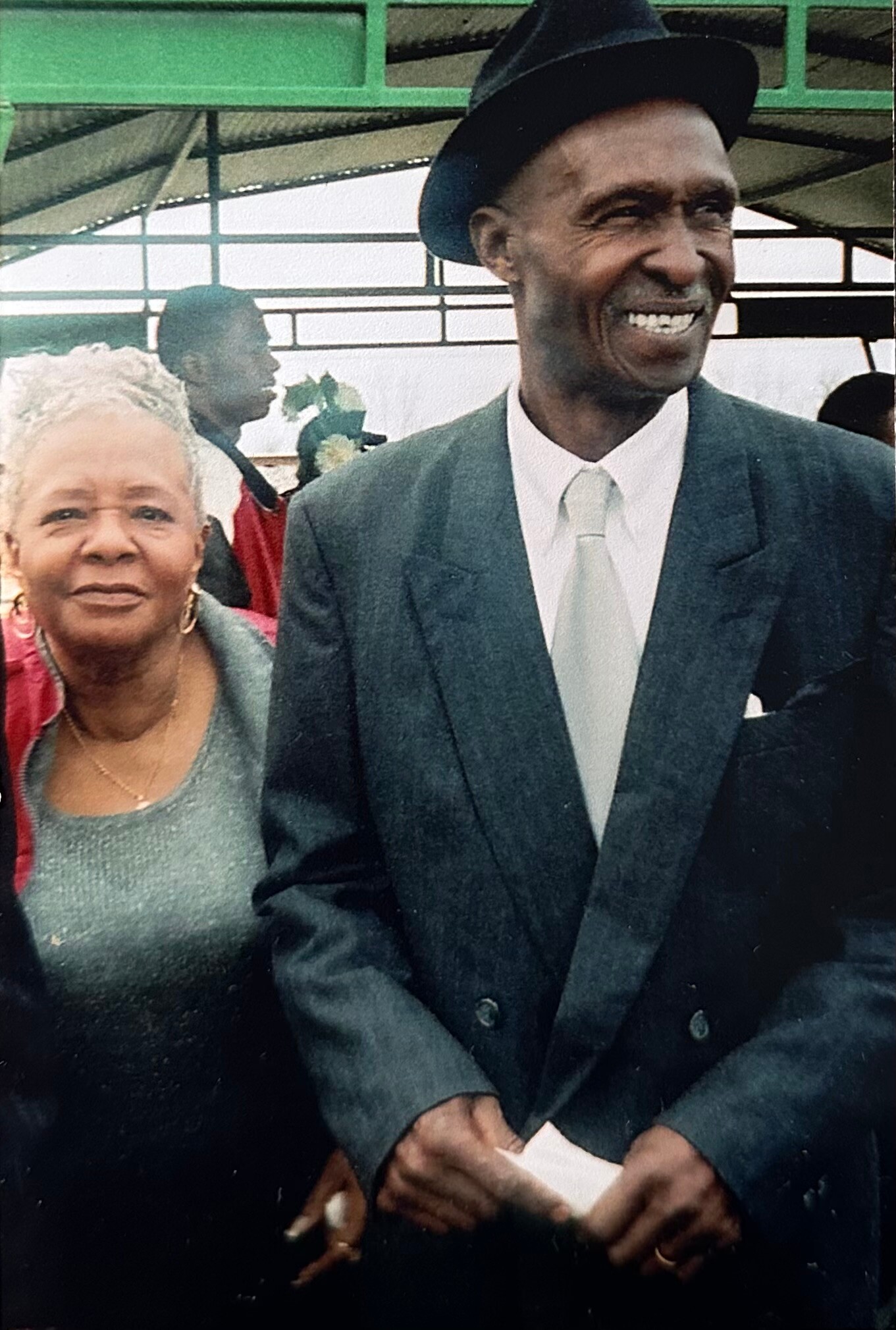 Smiling elderly man in a suit and fedora beside an elderly woman in a teal dress at a social event.