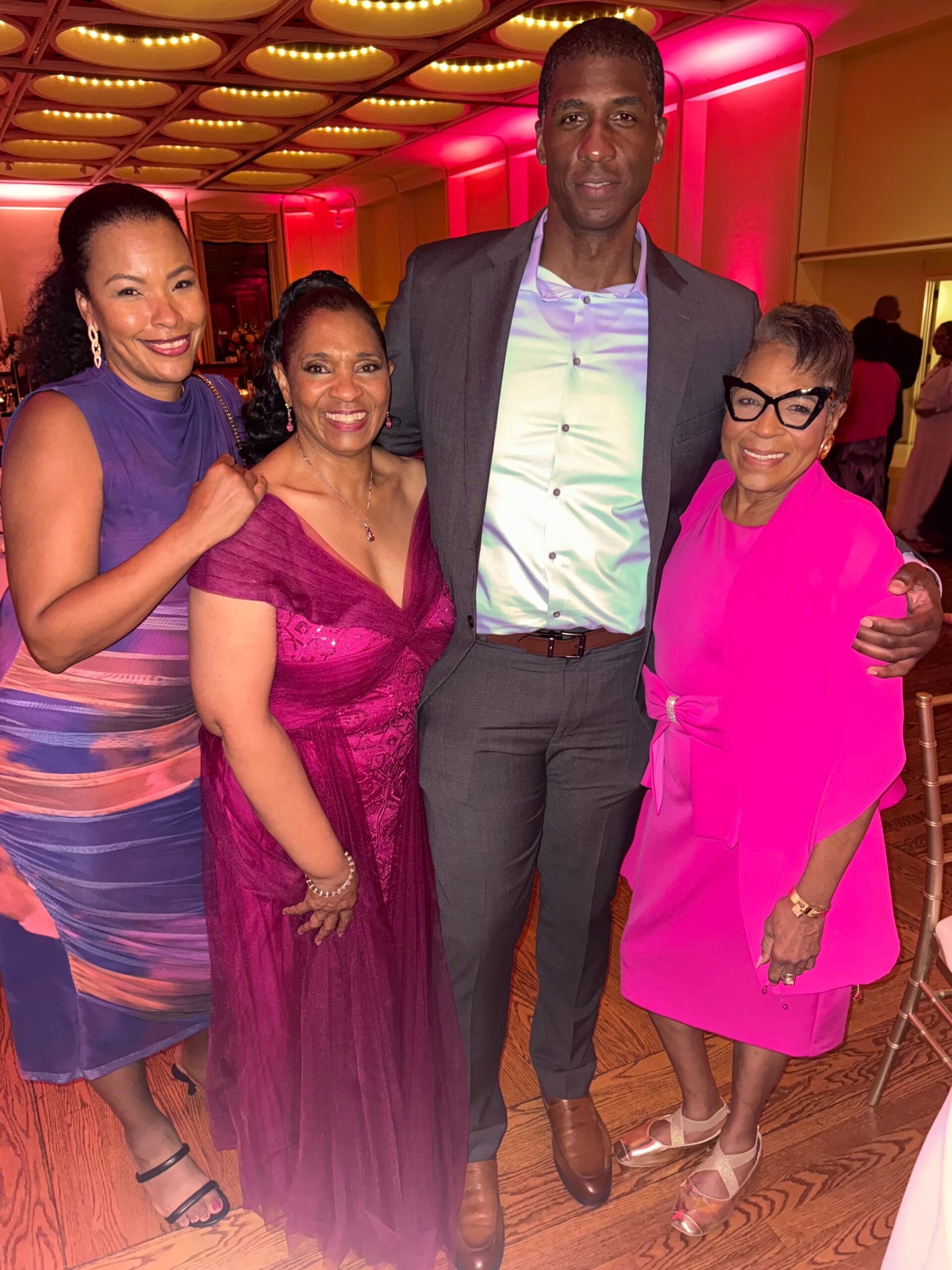 Four friends posing together at a formal event, smiling indoors under warm pink lighting; three women in vibrant dresses and one man in a suit.