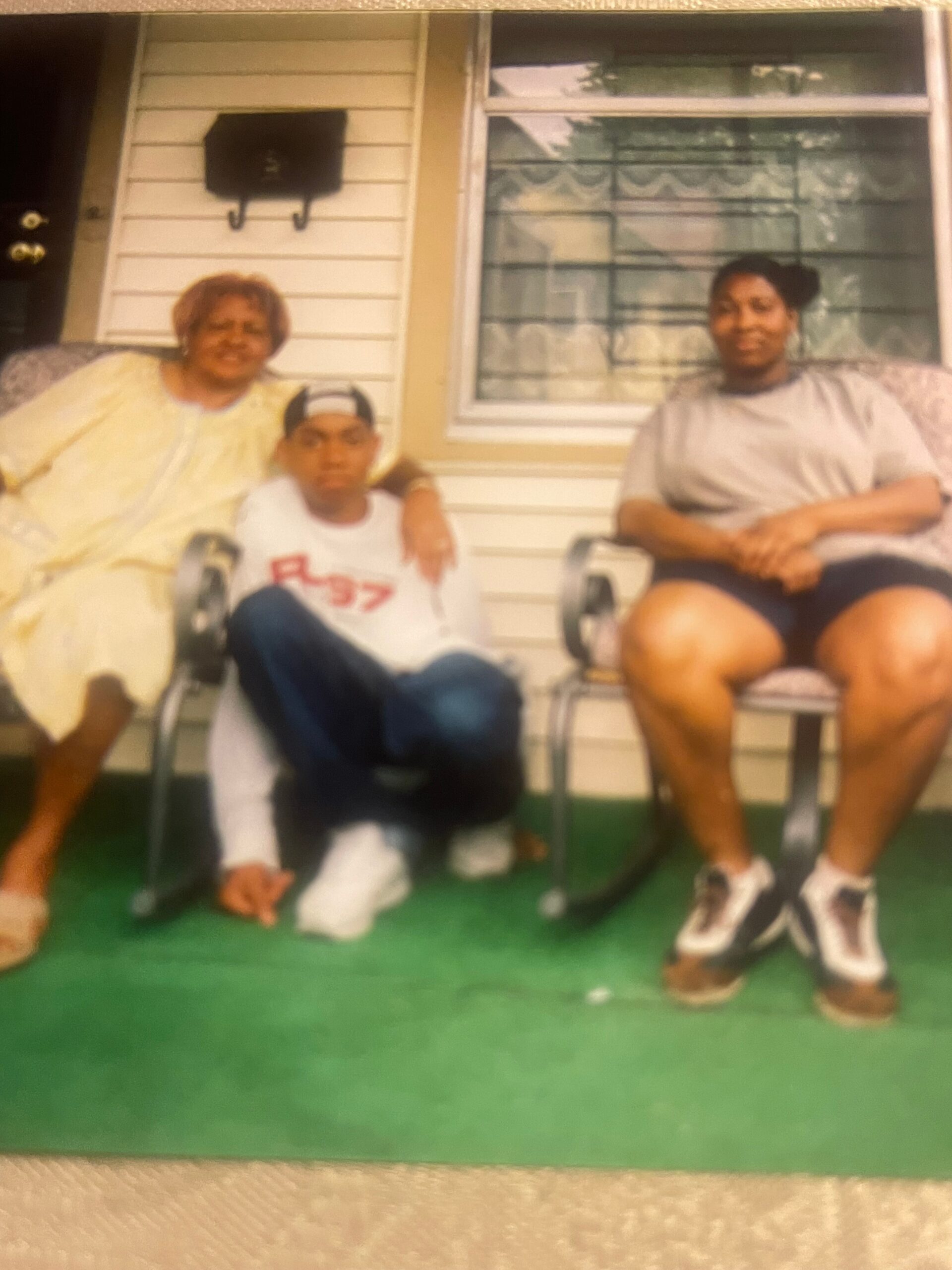 Three people sit together on a front porch: a woman in a yellow outfit on the left, a boy in a white shirt and backward cap in the middle, and a woman in gray on the right, with a window behind them.