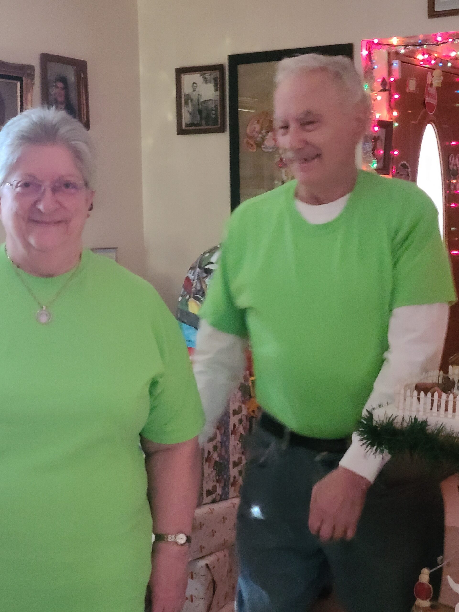 Smiling older couple wearing bright green shirts, standing indoors with festive holiday lights in the background.