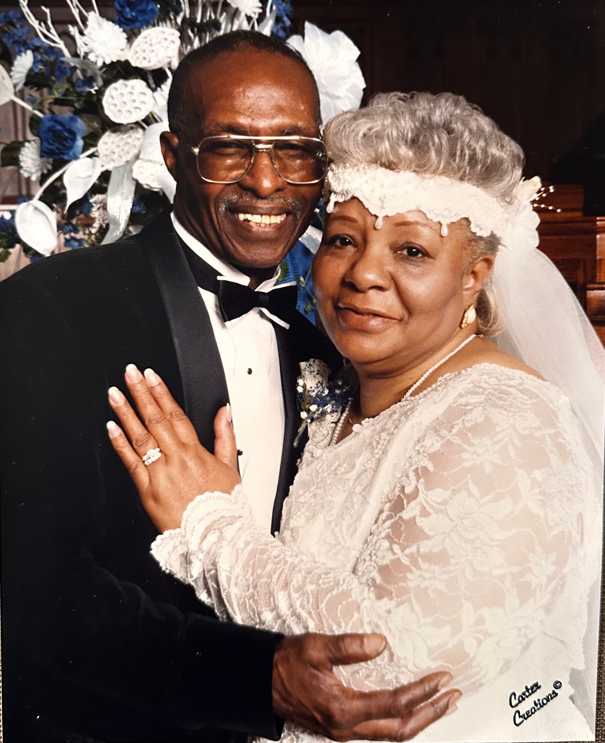 A smiling older couple at a wedding, the man in a tuxedo and bow tie embracing a bride in a lace dress with a floral background behind them.