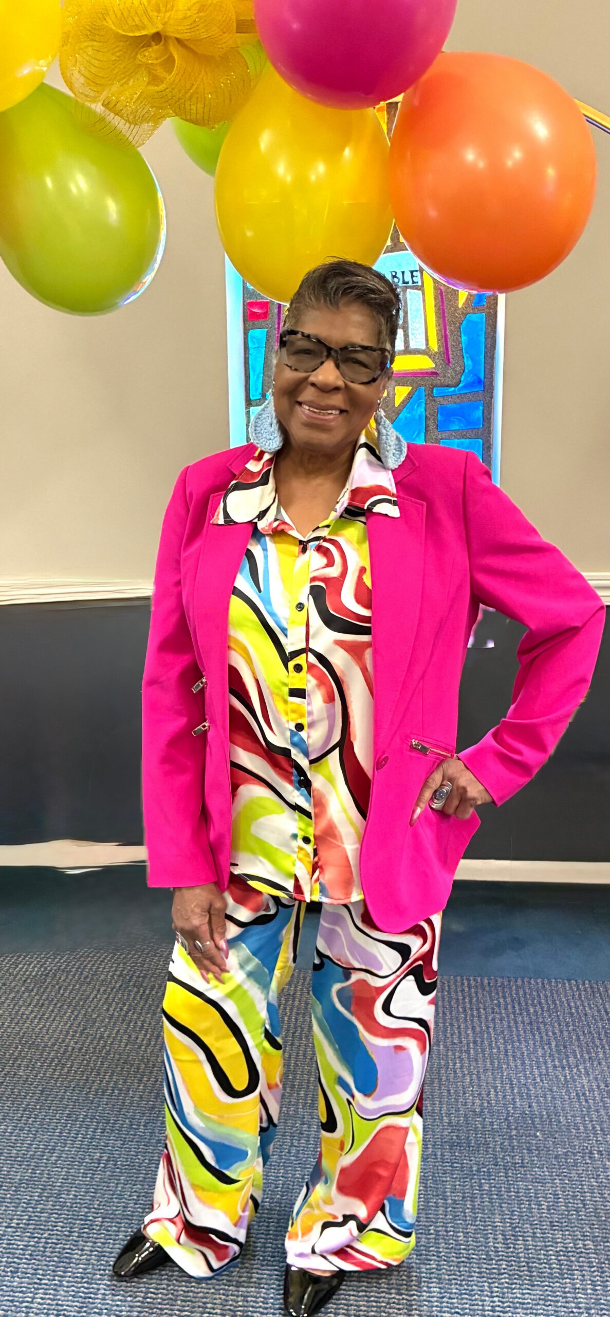 Smiling woman in a bright pink blazer and multicolored abstract outfit under colorful balloons at a celebration.