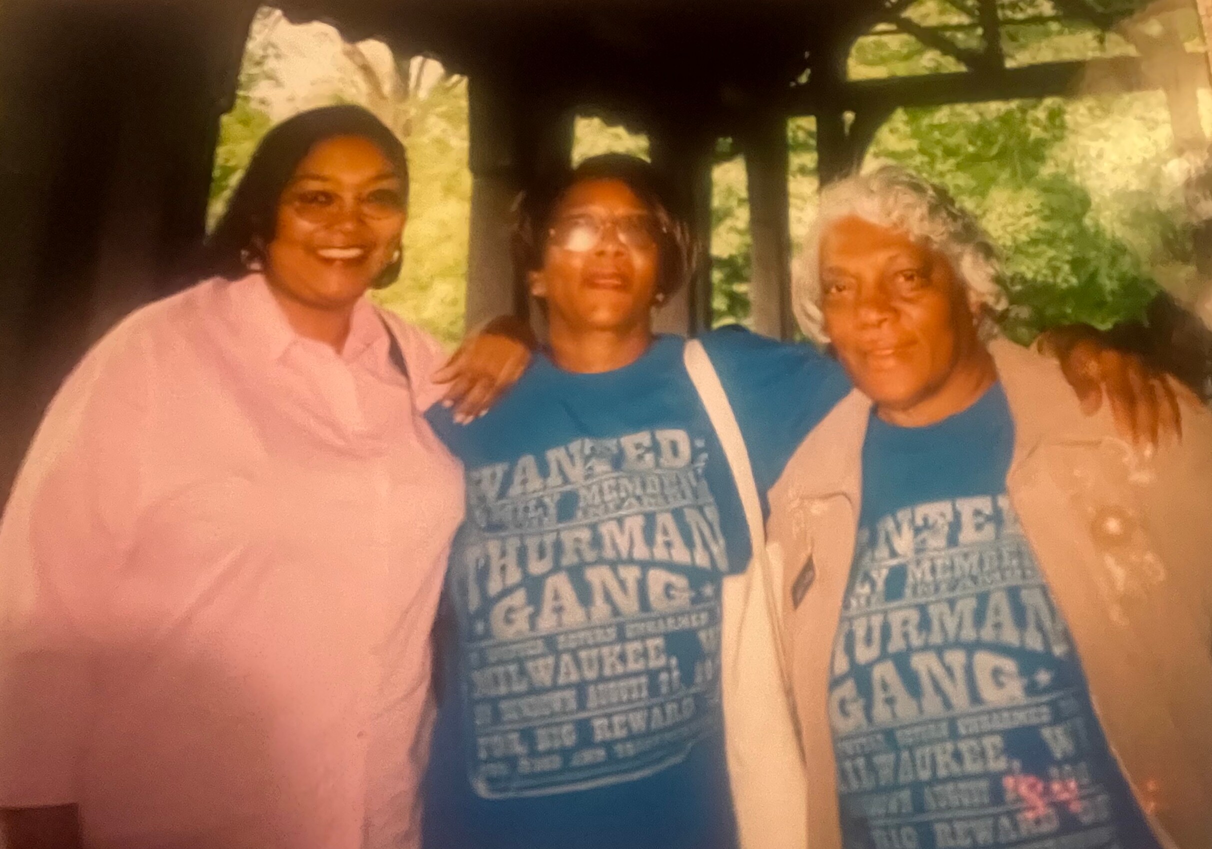 Three smiling women stand together in a shaded park gazebo, posing for a group photo.