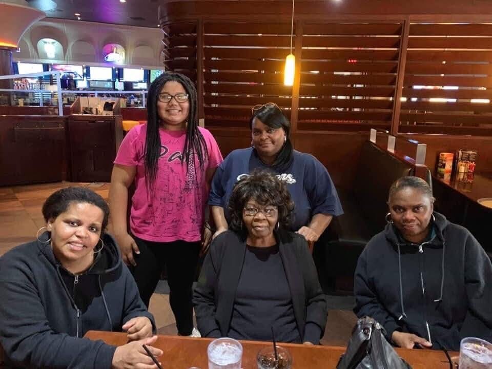 Five women pose for a group photo around a wooden restaurant table, some seated and some standing, smiling at the camera.