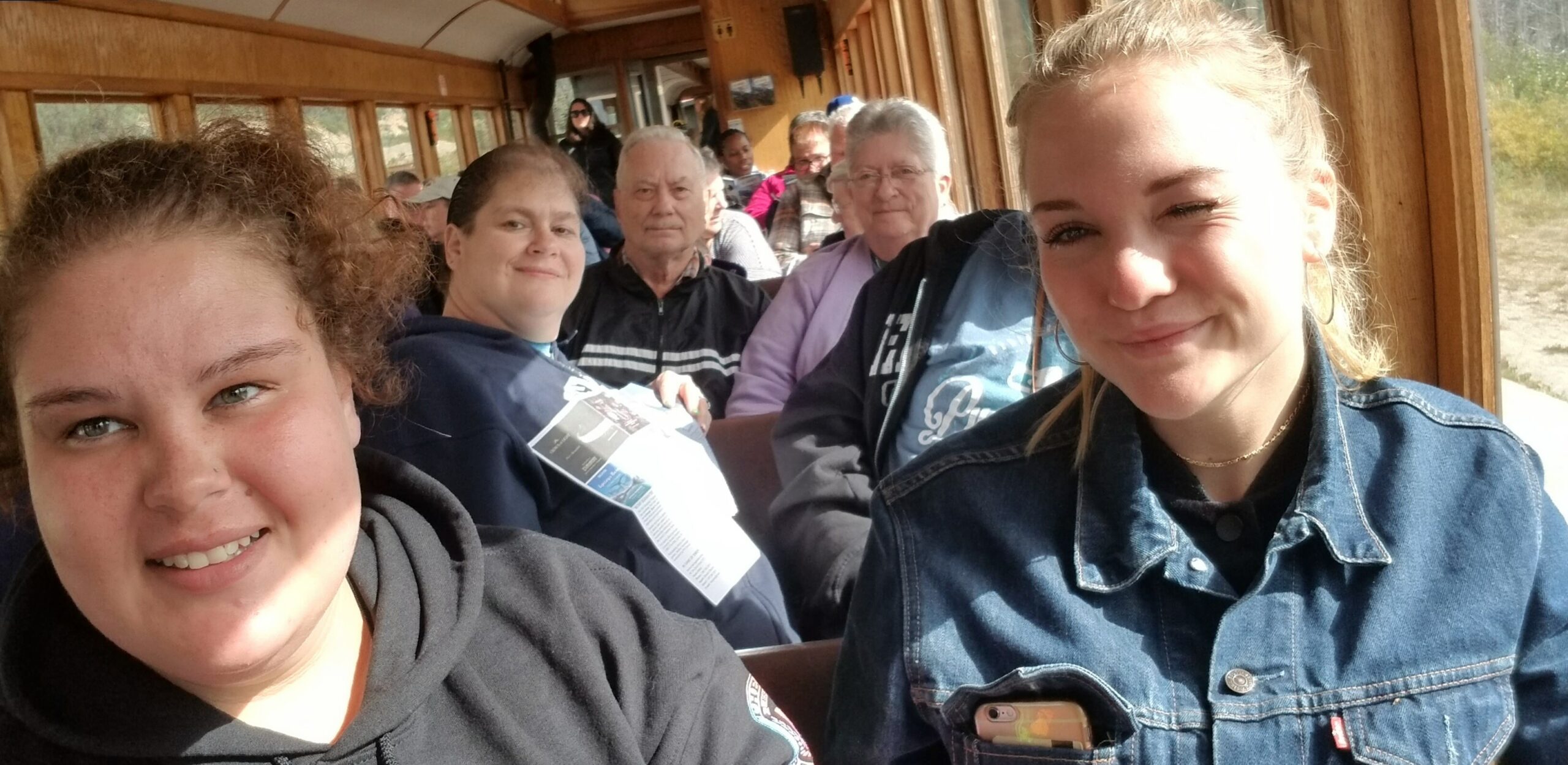 Two young women in the foreground smiling for a selfie on a wooden-train style passenger vehicle, with other riders behind them.