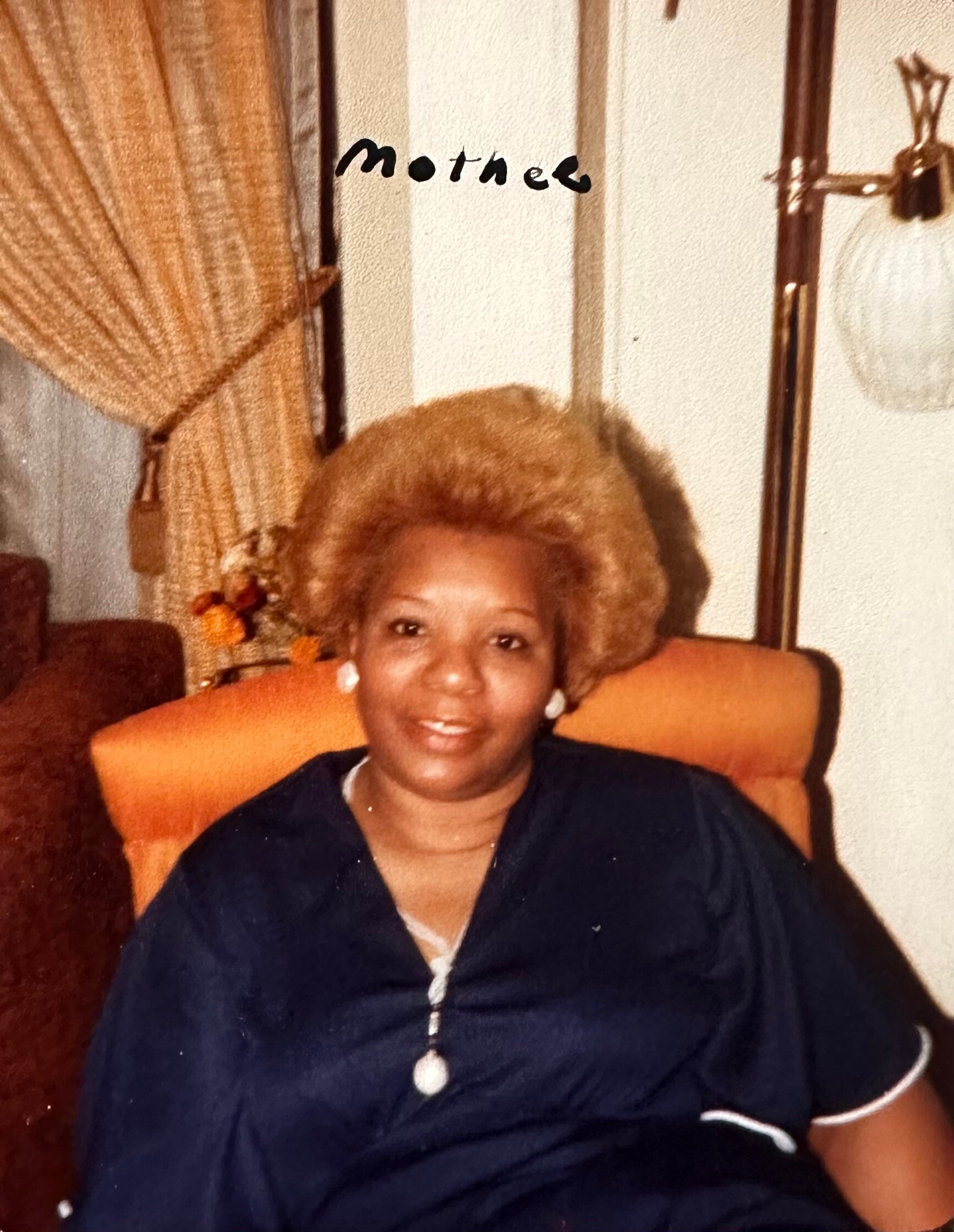 Smiling woman seated in an orange chair wearing a dark top and pendant necklace, with beige curtains and a lamp in the background.