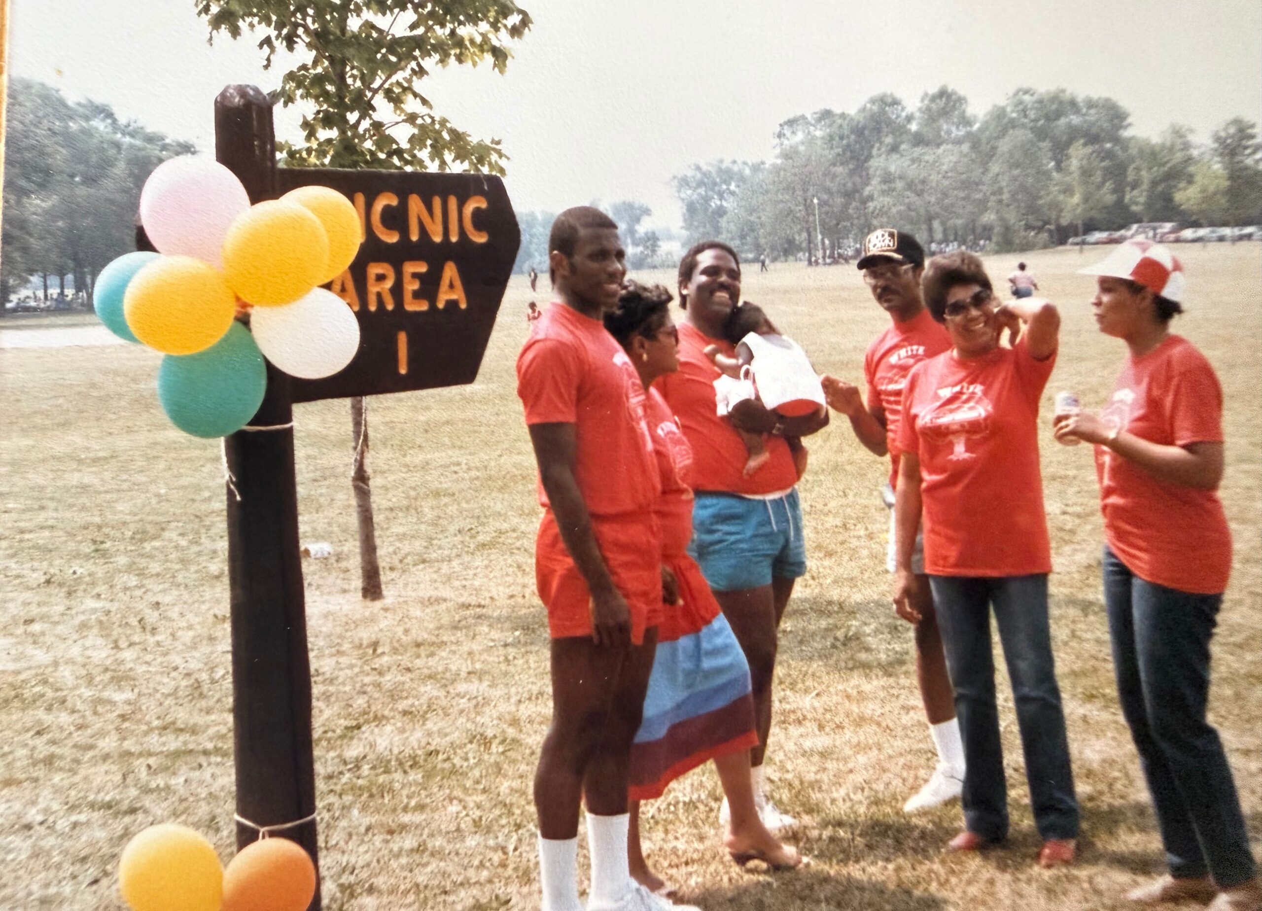 Group of friends in red shirts stand in a park beside a wooden post with colorful balloons and a 'Picnic Area' sign.