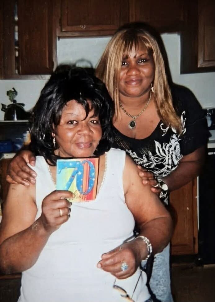 Two women in a kitchen posing; the seated woman displays a colorful '70' birthday card while smiling, with a friend standing behind her.