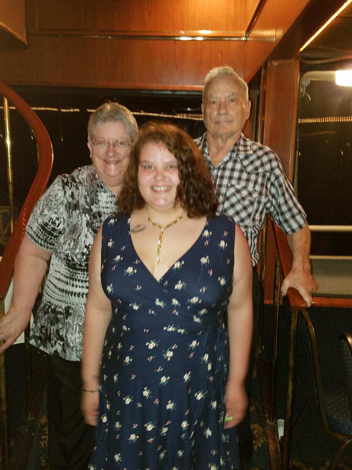 Three people pose on a stairway: a smiling young woman in a navy floral dress at the front, with an older woman and man behind her.