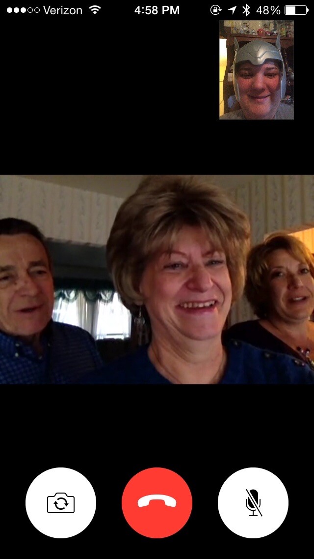 Group video call on a phone: three smiling people in a living room, with a small inset video of a person wearing a helmet costume in the top-right; bottom controls show camera, end call, and mute buttons.