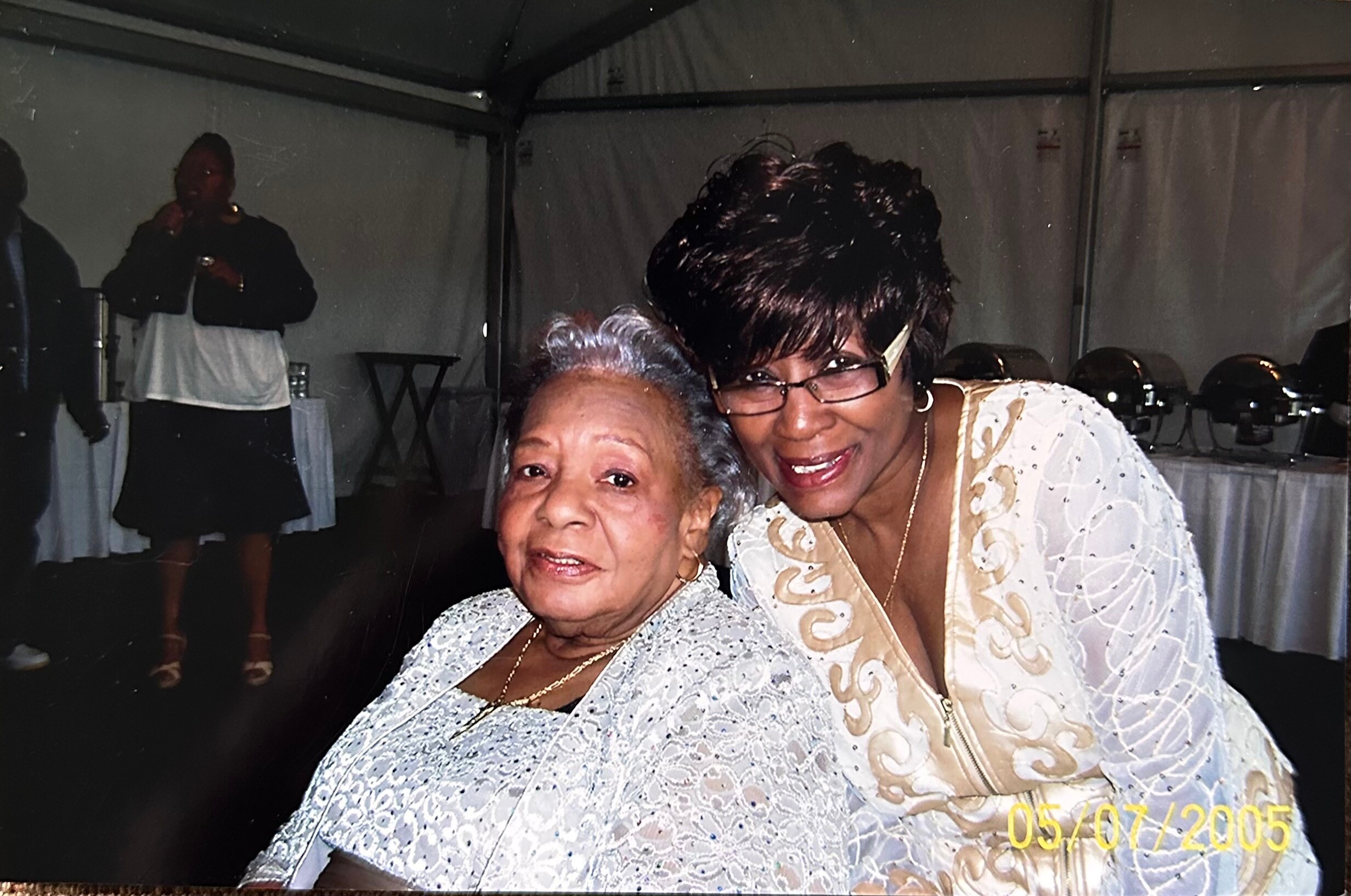 Two smiling women posing closely together at a celebration, wearing light-colored outfits inside a decorated venue.