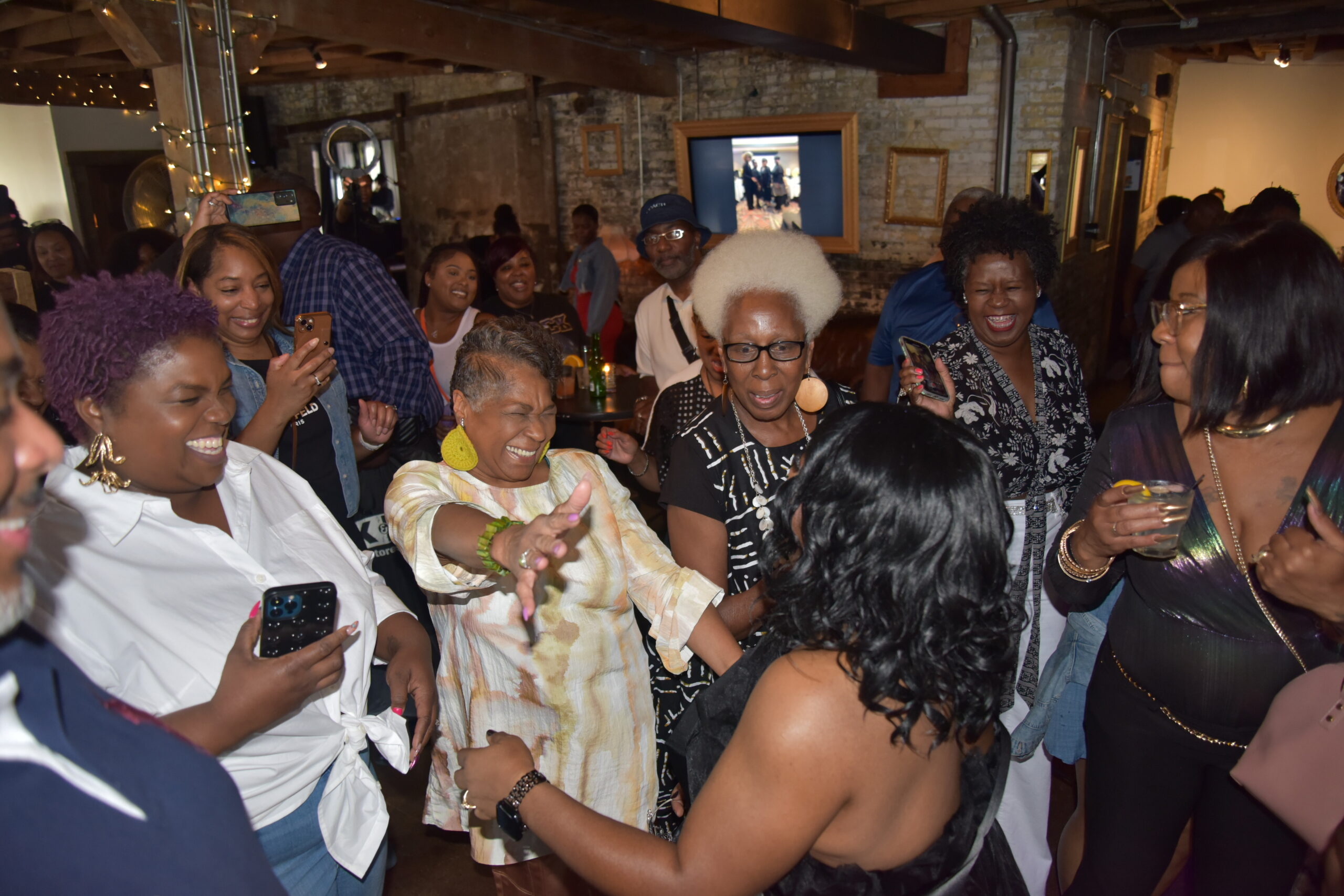 People dancing and smiling at a crowded indoor party with exposed brick walls and string lights in the background.