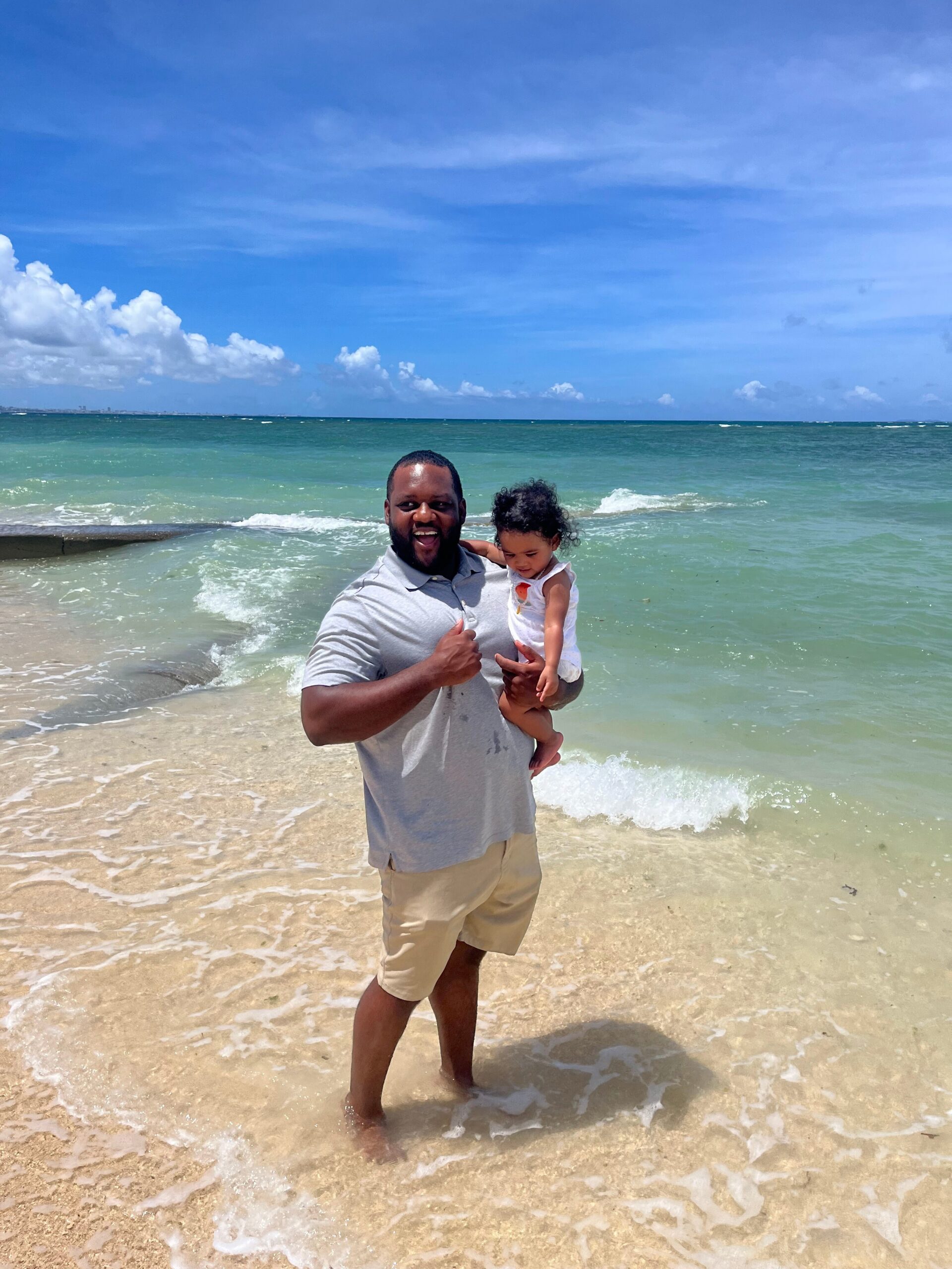 Man holding a small child on a sunny beach, standing in shallow water as waves roll in.