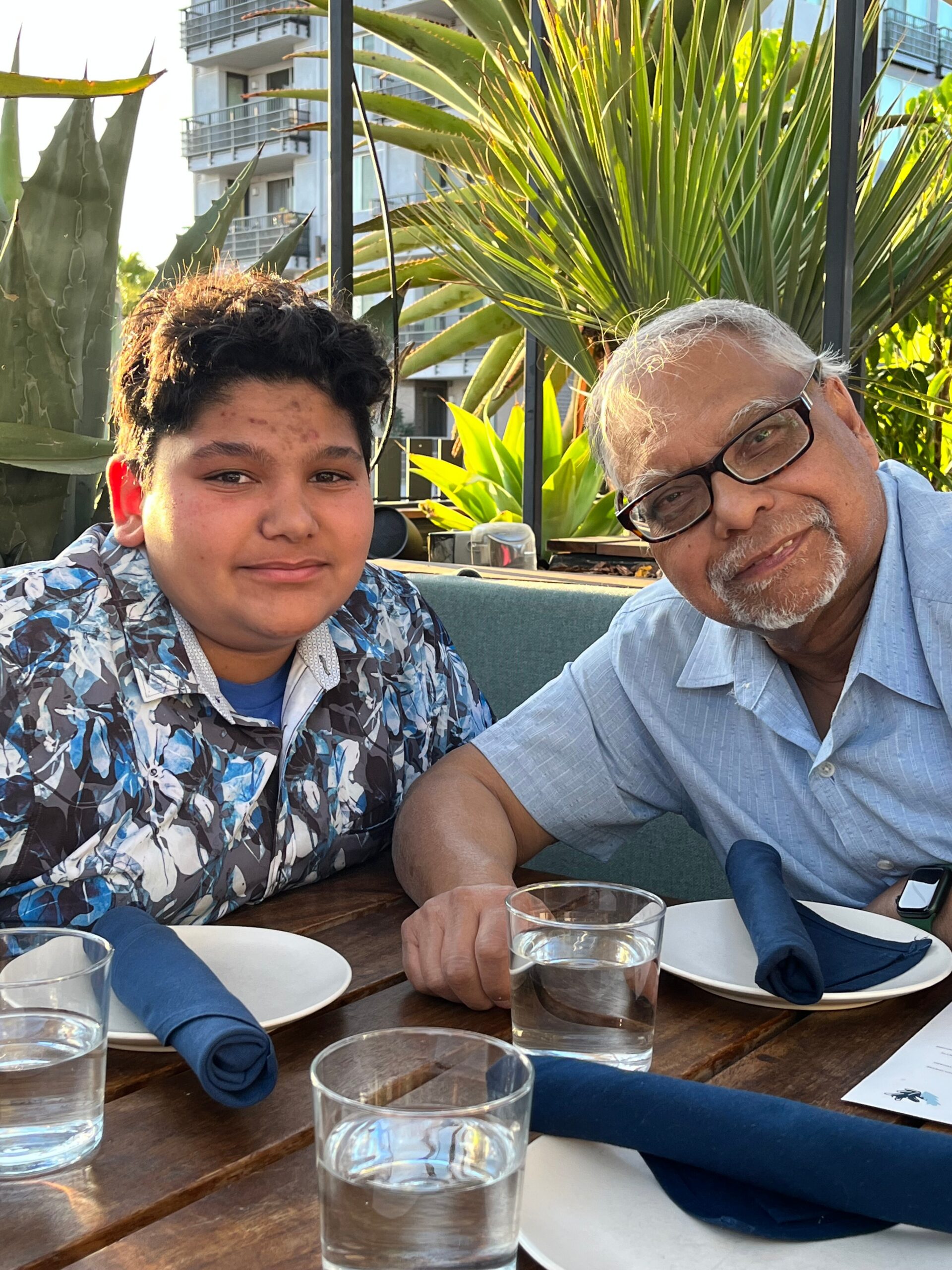 Two men sitting at an outdoor dining table with blue napkins, smiling, and glasses of water in front; greenery and modern buildings in the background.