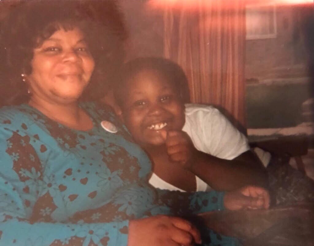 Smiling woman in a teal floral dress sitting with a boy giving a thumbs up indoors.