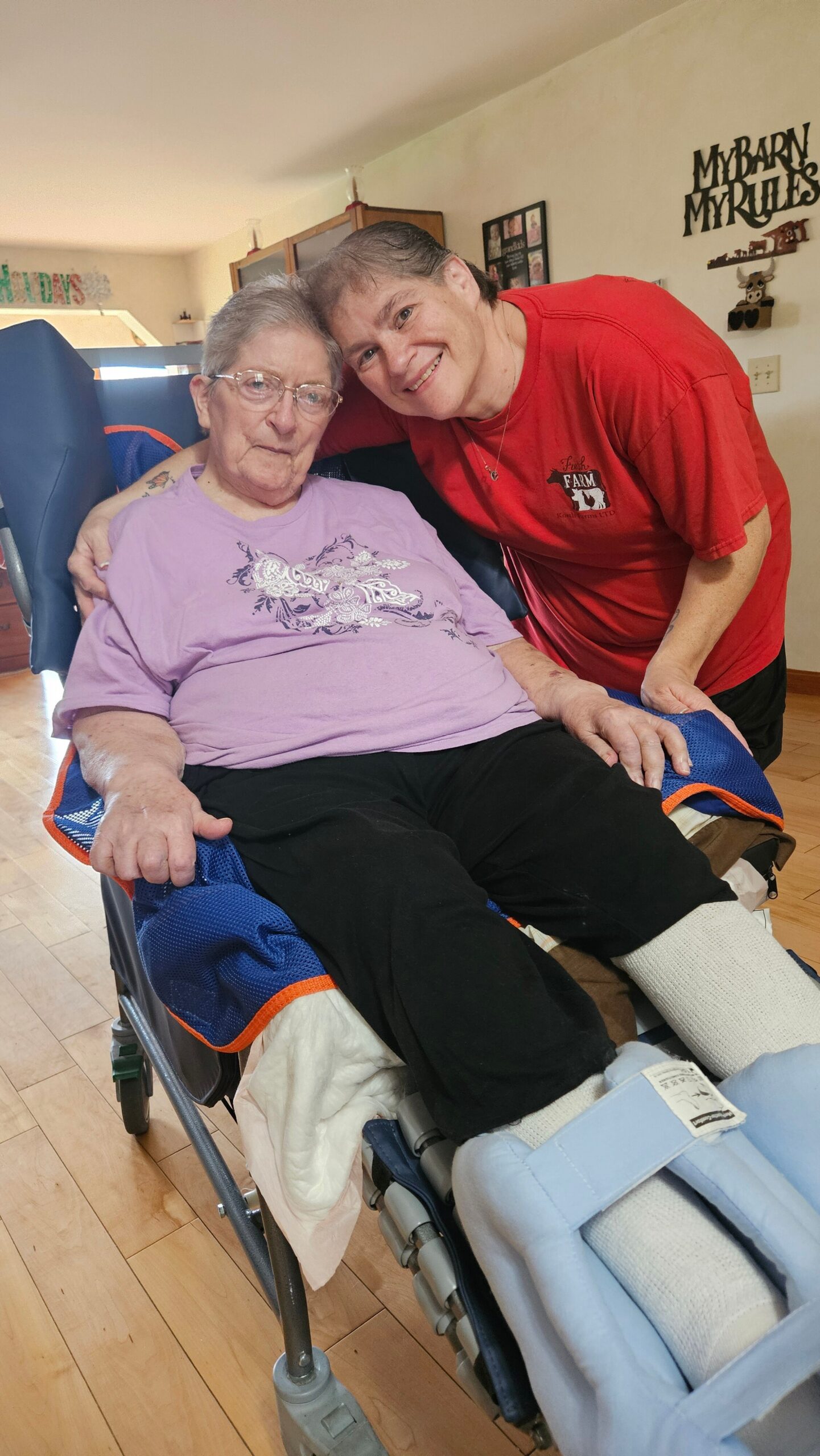 Caregiver standing beside an elderly woman in a purple shirt who sits in a wheelchair, both smiling at the camera.