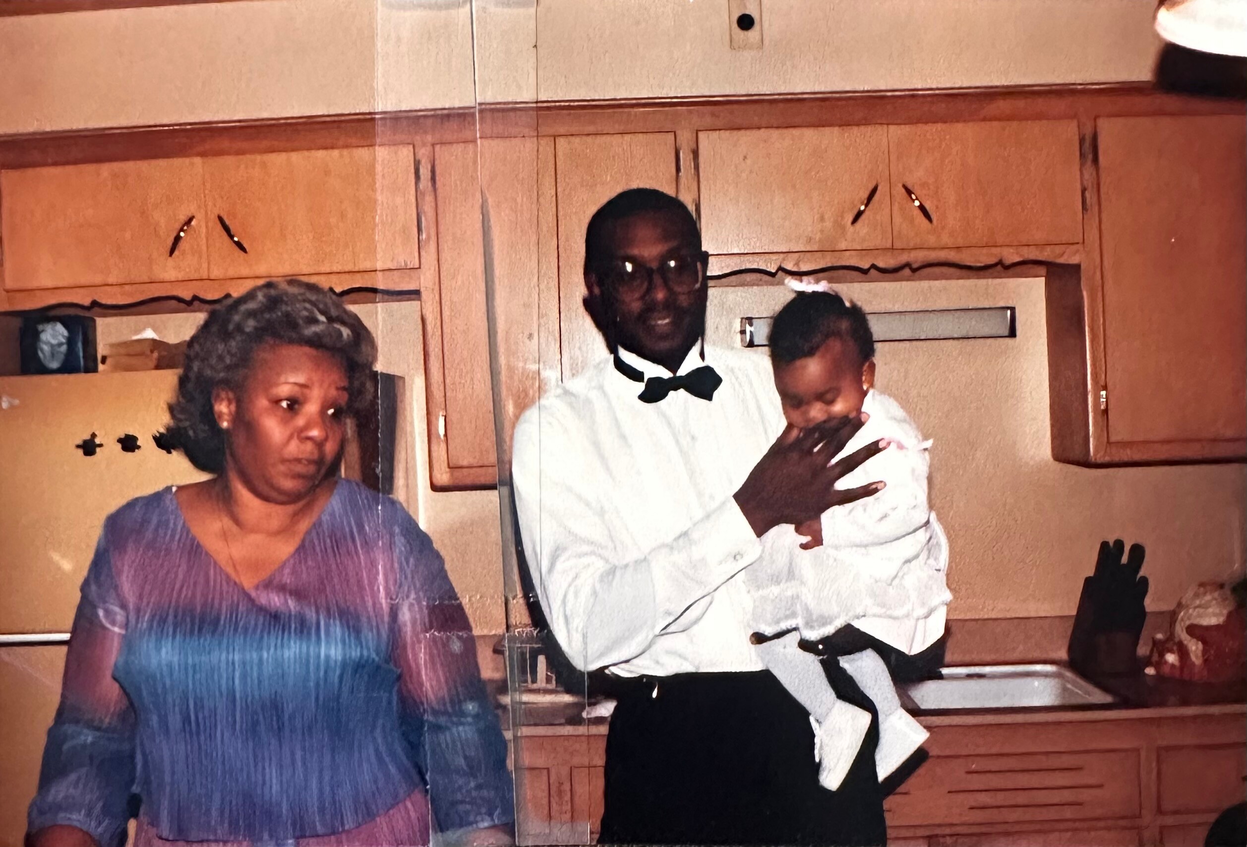 Woman in a purple blouse stands beside a man in a white shirt and bow tie who is holding a baby in a kitchen with wooden cabinets behind them, a family portrait.
