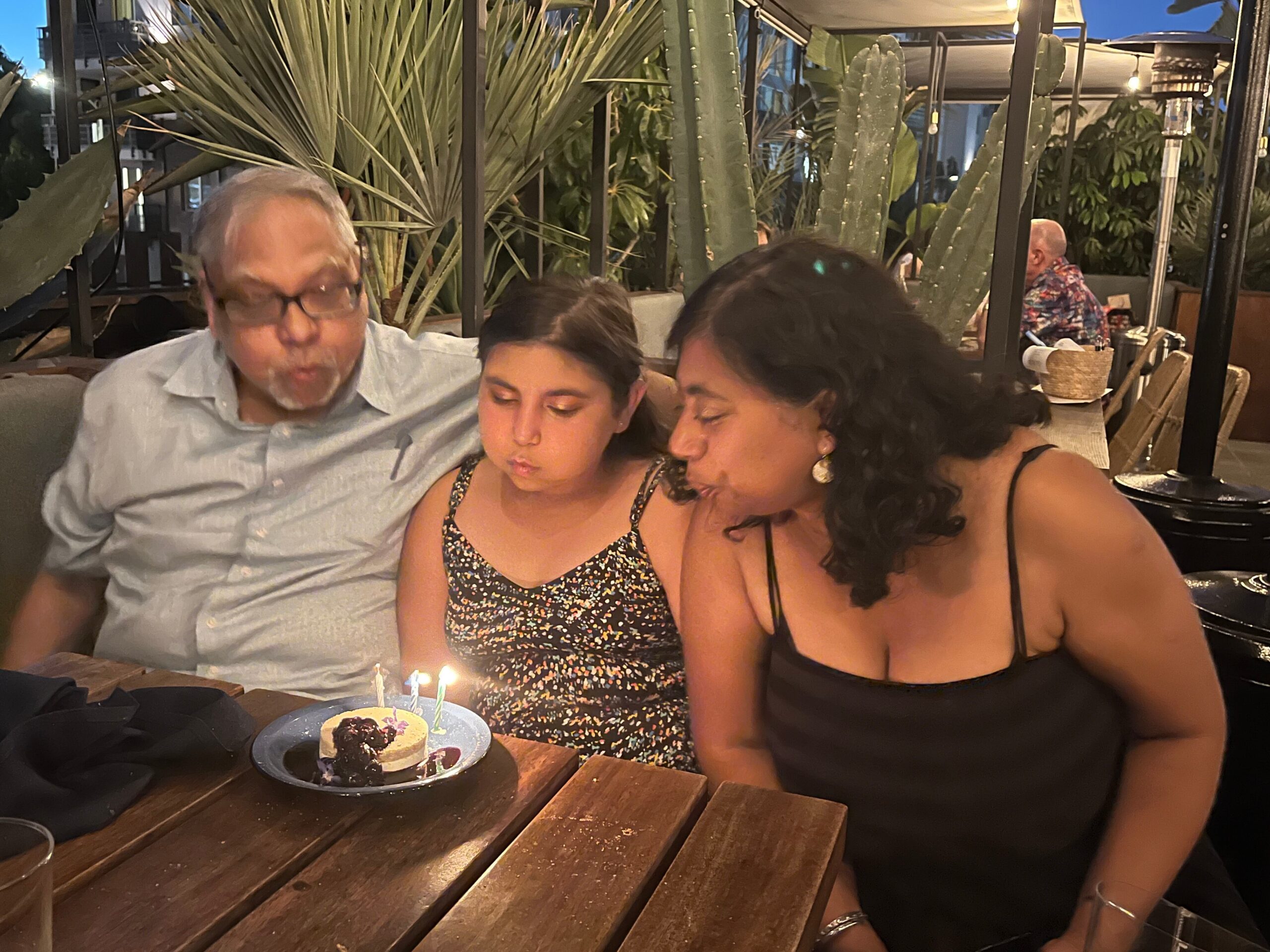 Group of three people at a wooden table celebrating a birthday, cake with lit candles in front of them, outdoor plant setting in evening light.