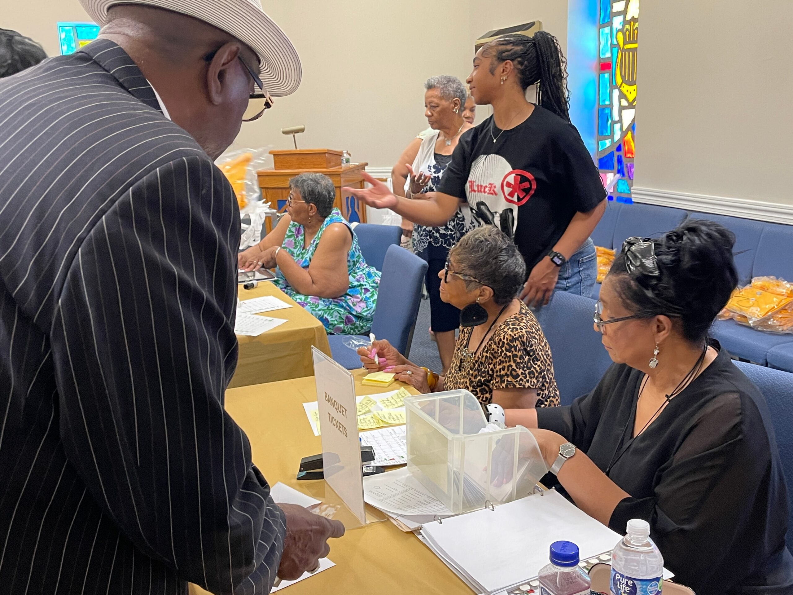Attendees at a banquet-style check-in table; a woman in black helps guests as others sit and fill forms in a church hall.