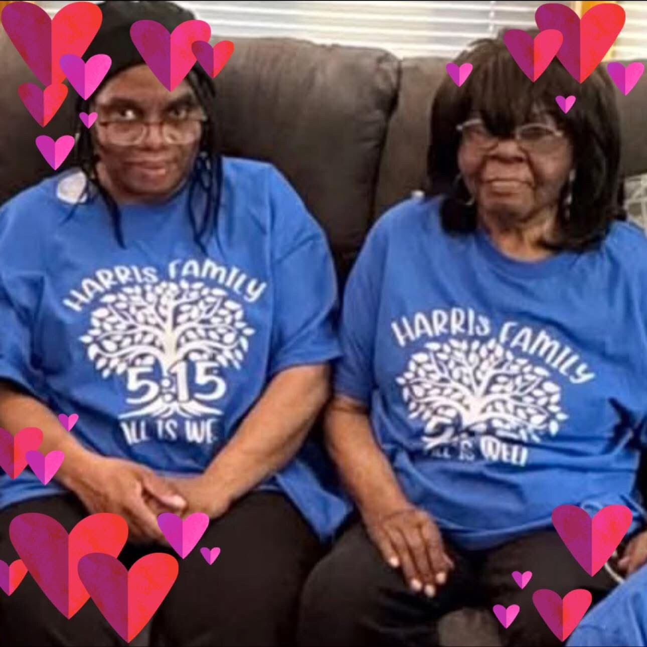 Two women in matching blue Harris Family shirts sit on a couch, hand in hand, with pink and red heart decorations around them.