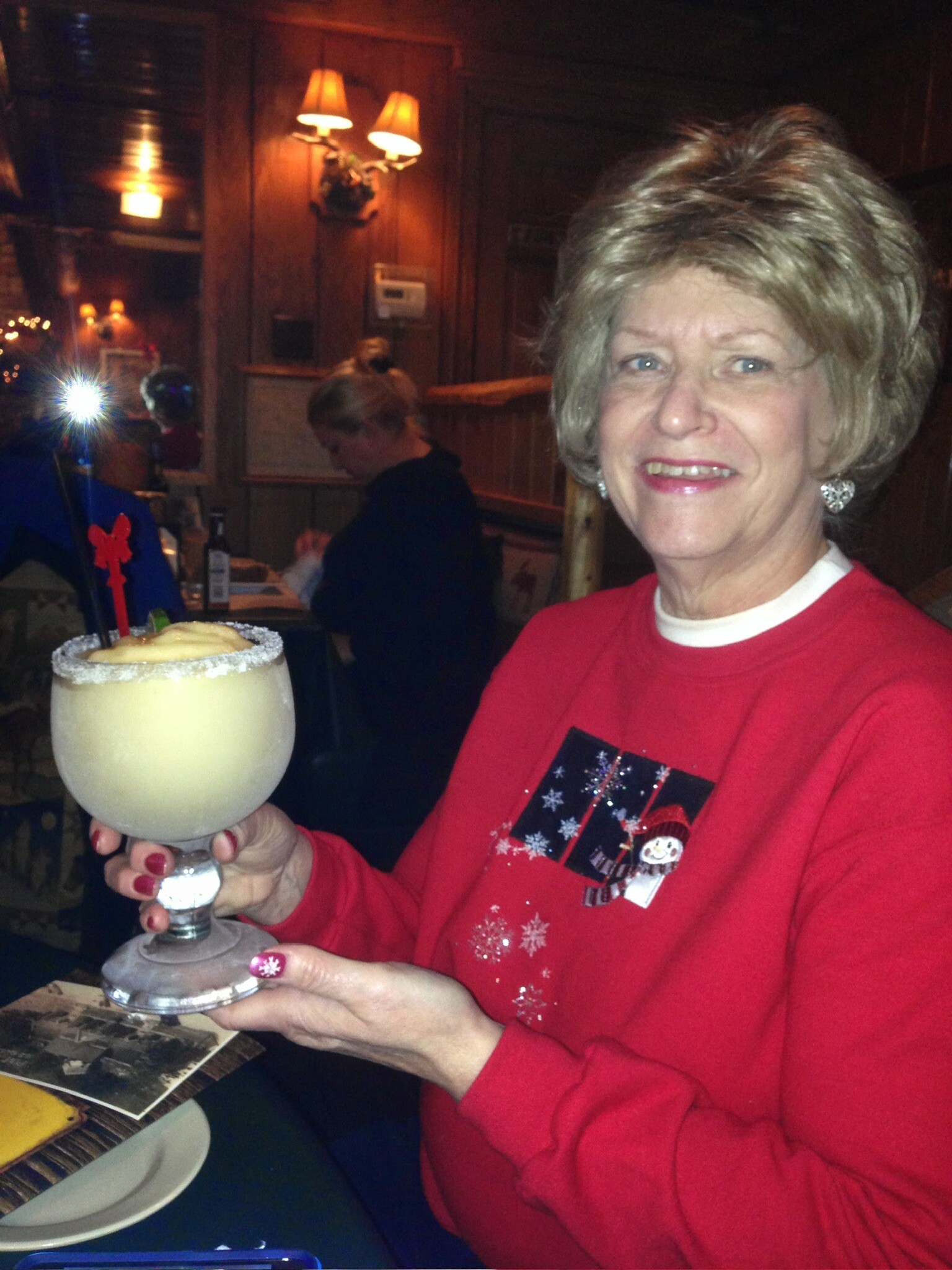 Smiling woman in a red sweater holding a large salted-rrim cocktail in a dimly lit bar/restaurant.