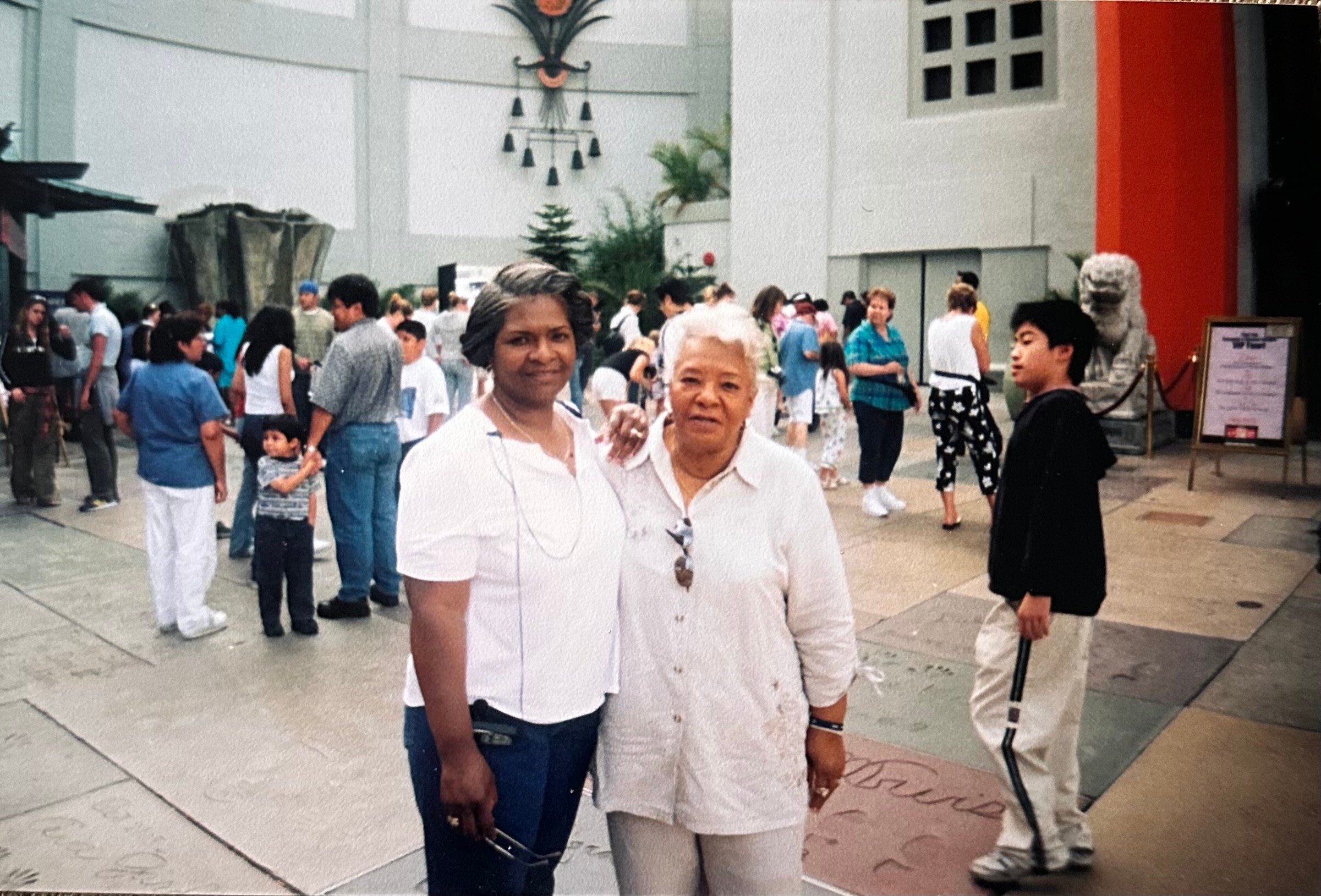 Two women in white shirts stand close together, smiling, with a crowded outdoor plaza behind them at a public event.