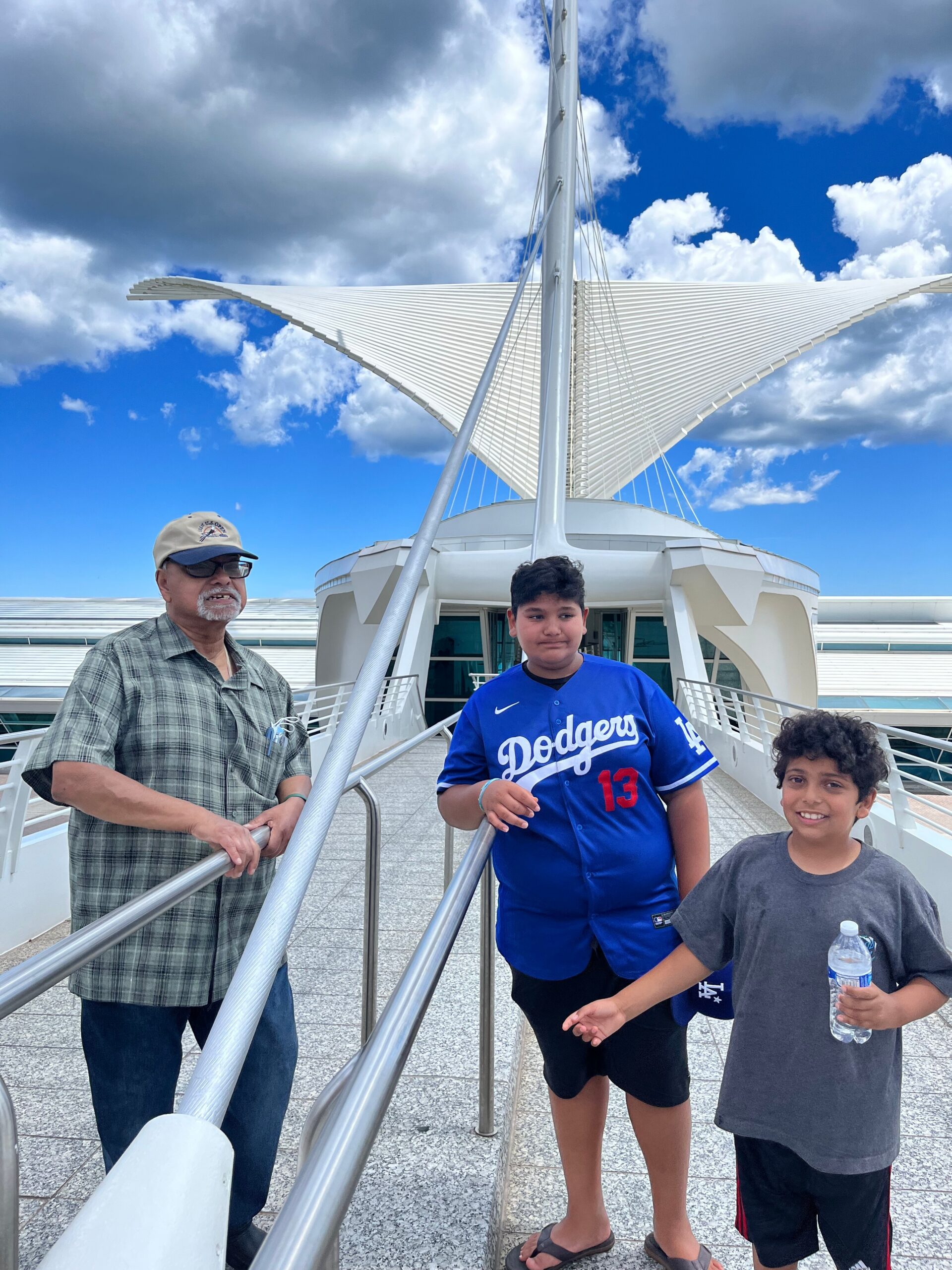 Three people stand on a ship deck next to a tall white mast with a wing-like sail structure against a partly cloudy blue sky.