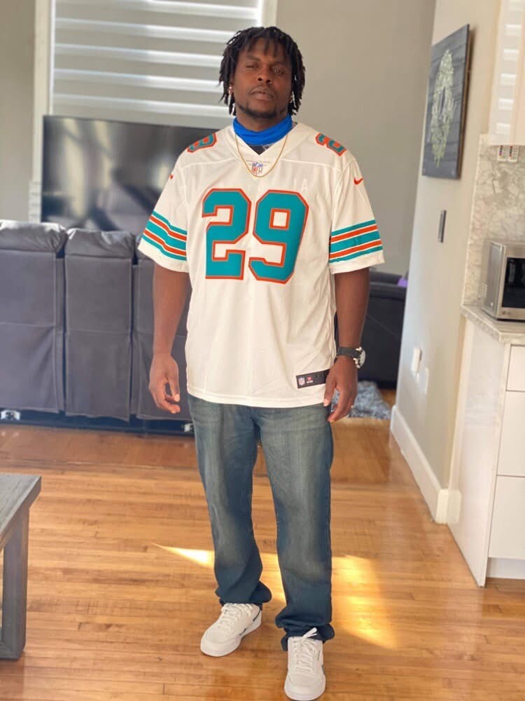 Man standing in a living room wearing a white Miami Dolphins jersey with teal/orange numbers (#29), jeans, and white sneakers.