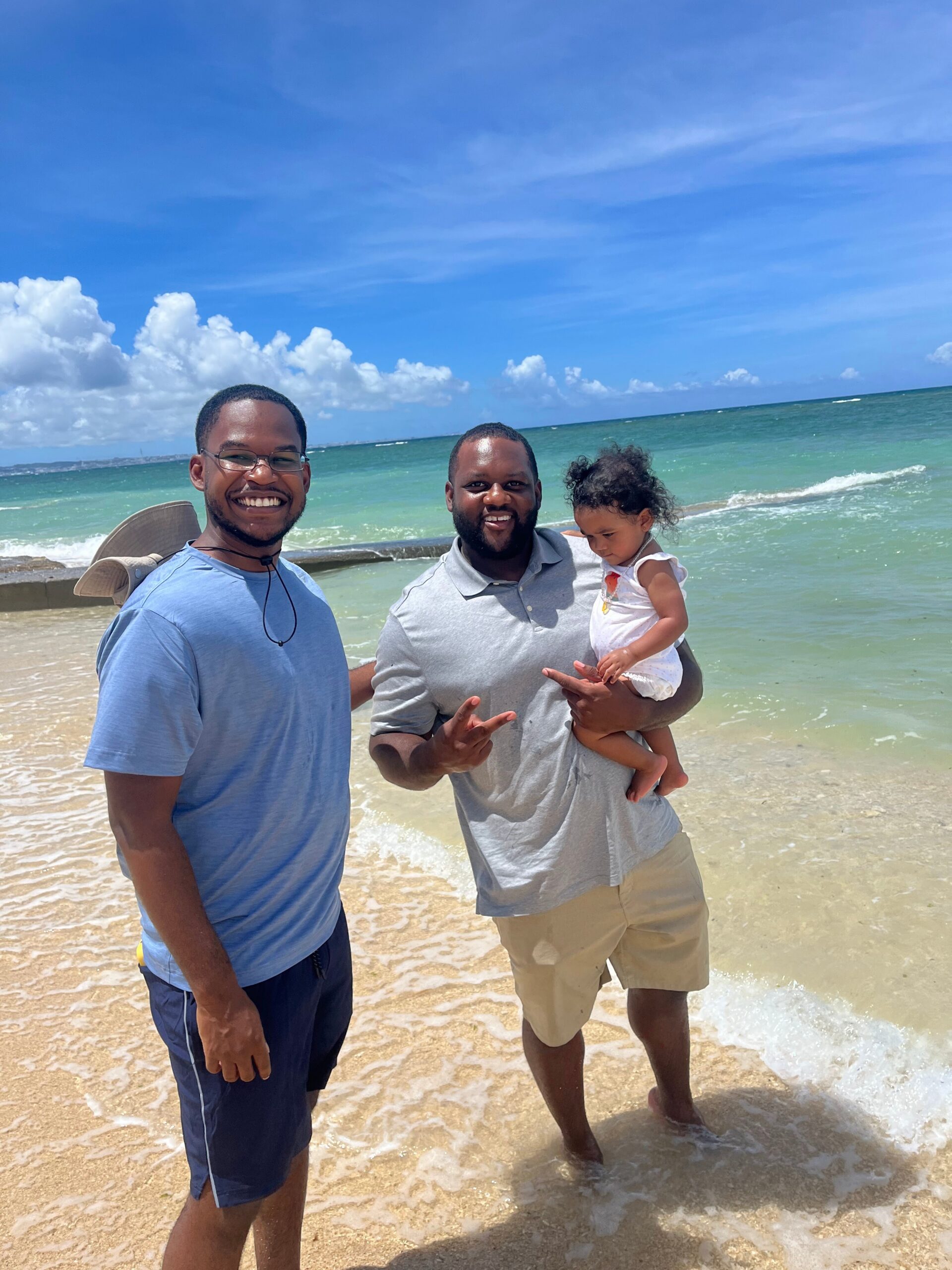 Three people at the beach posing for a photo: two men smiling, one holding a small child, with waves and blue sky behind them.
