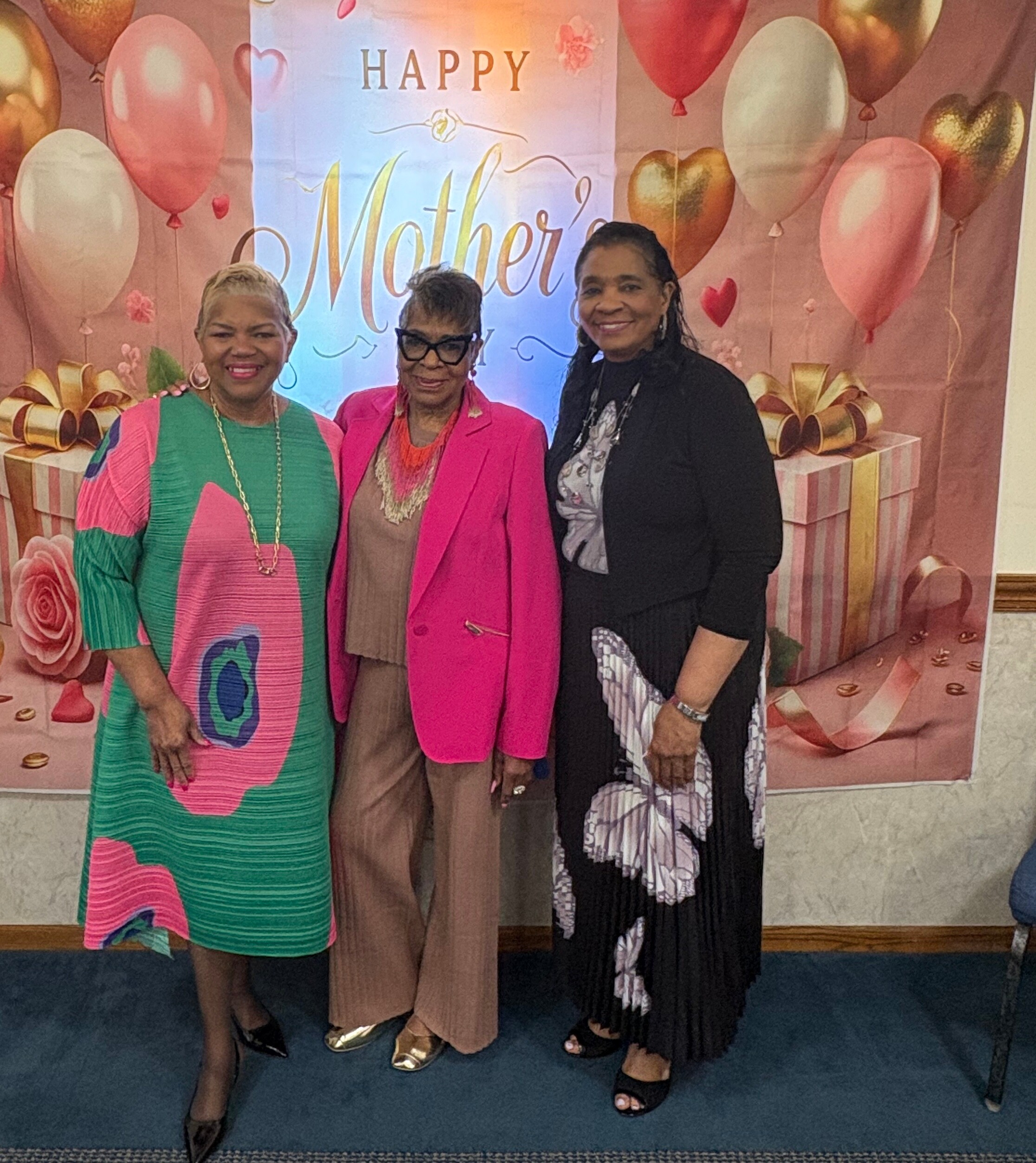 Three women posing in front of a Mother's Day backdrop with balloons and gifts, smiling.
