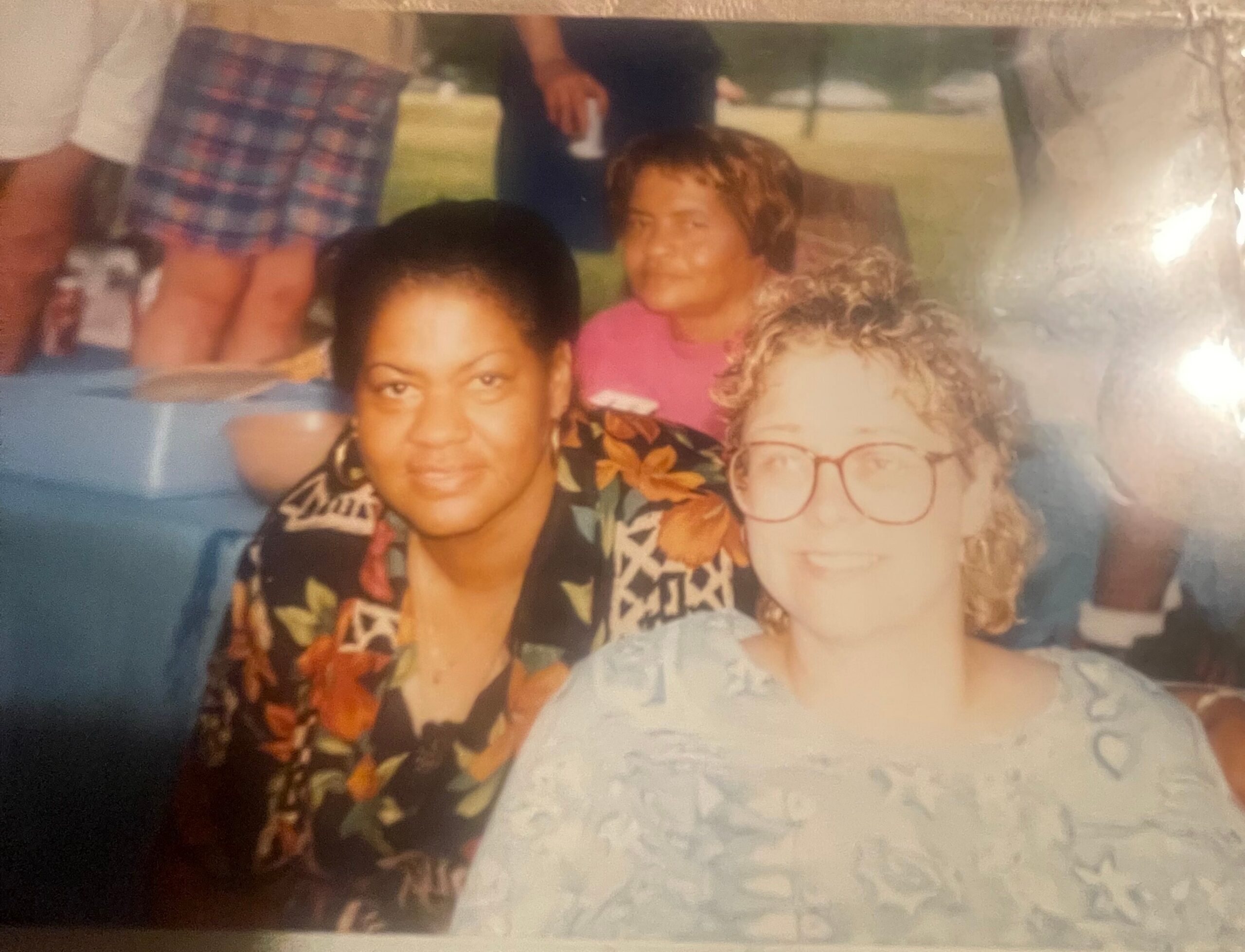 Three women posing for a photo outdoors; woman with dark hair and floral shirt in front, woman with glasses at right, and a person in a pink shirt in the background.