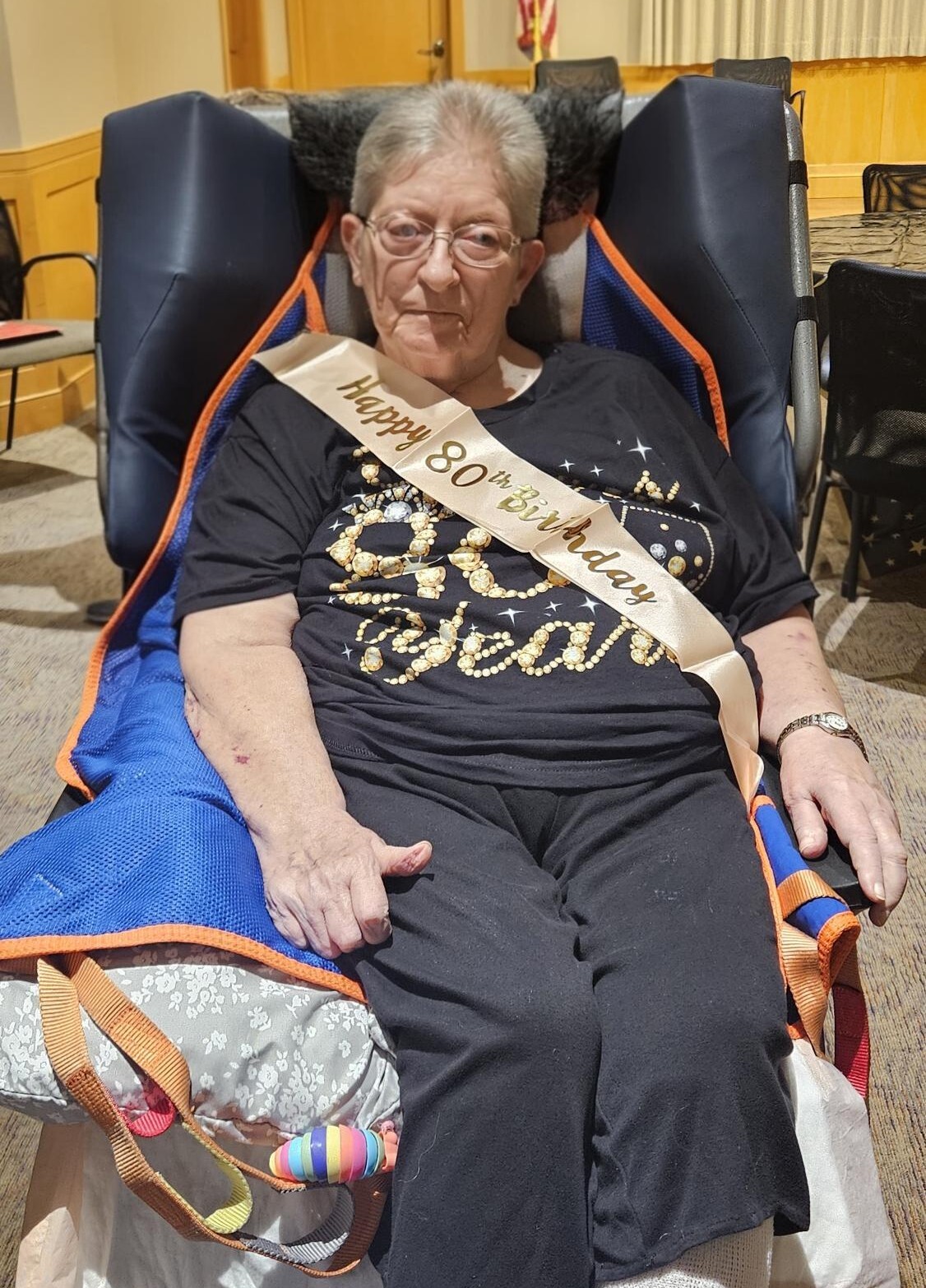 Older woman wearing a 'Happy 80th Birthday' sash sits in a blue-orange cushioned chair at a celebration.