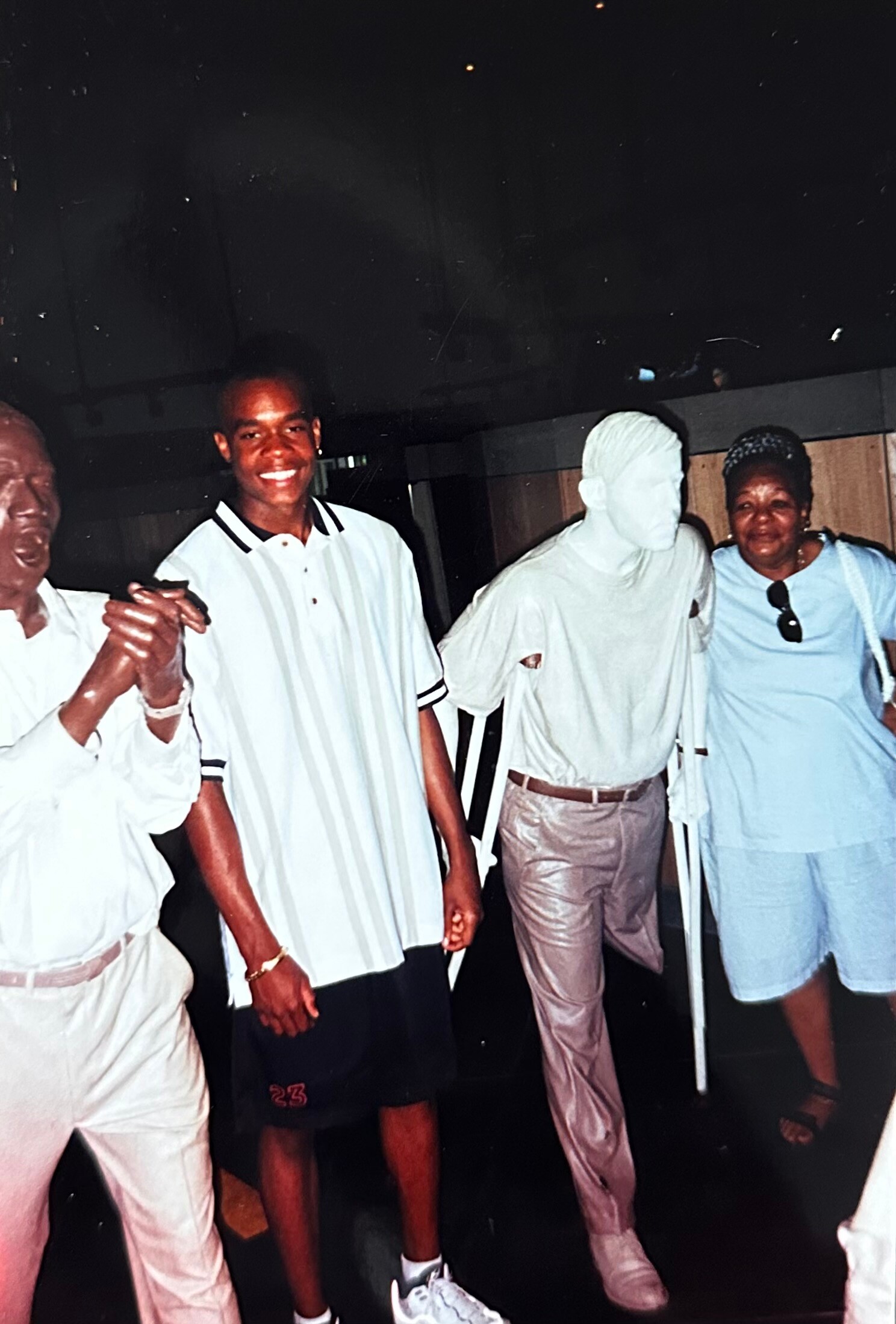 Four people posing together and smiling, holding hands in a casual indoor setting, wearing light-colored outfits.