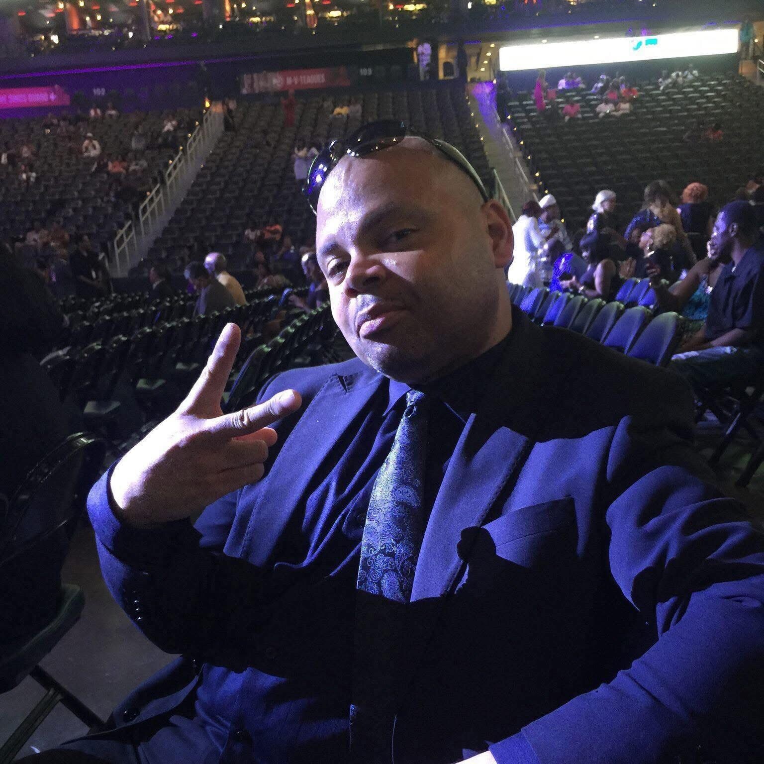 Man in a blue suit and tie taking a selfie at a large indoor arena, flashing a peace sign.