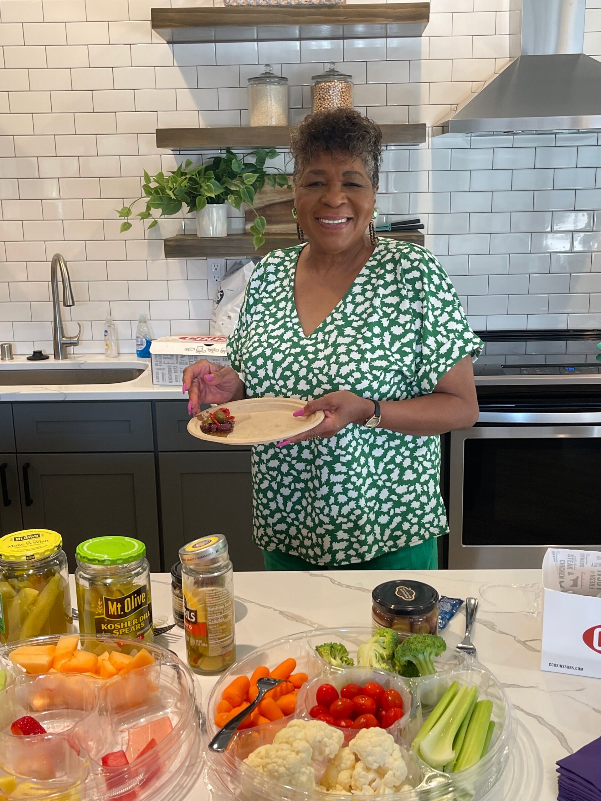 Smiling woman in a green patterned dress stands in a bright kitchen, holding a plate of food.