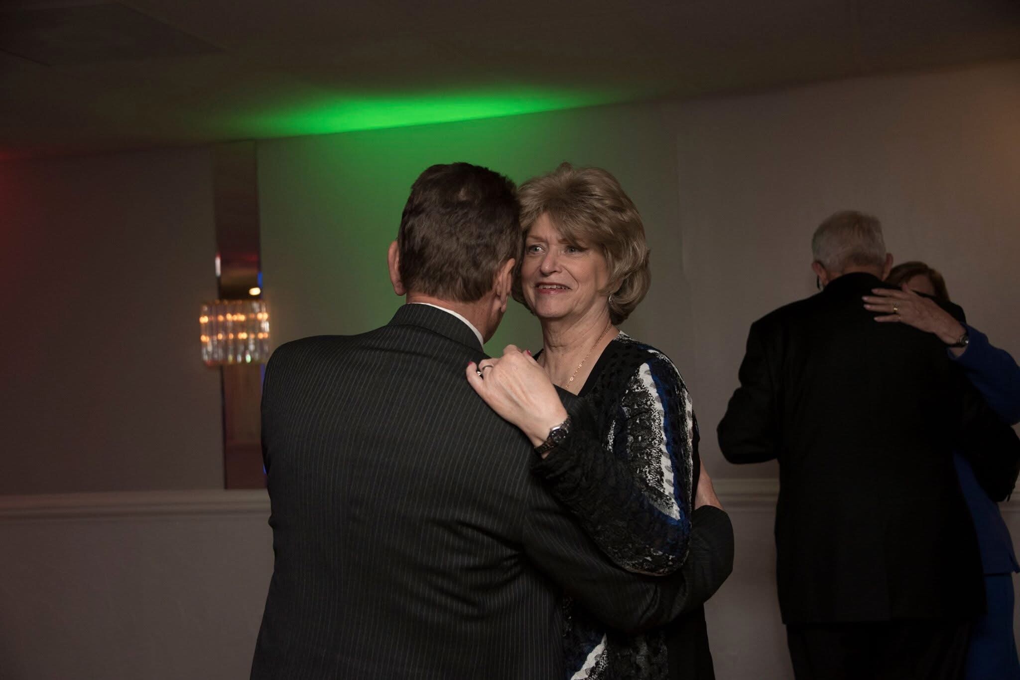 Couple dancing closely in foreground at a dimly lit event, woman looking at her partner while other couple hugs in the background with green lighting overhead