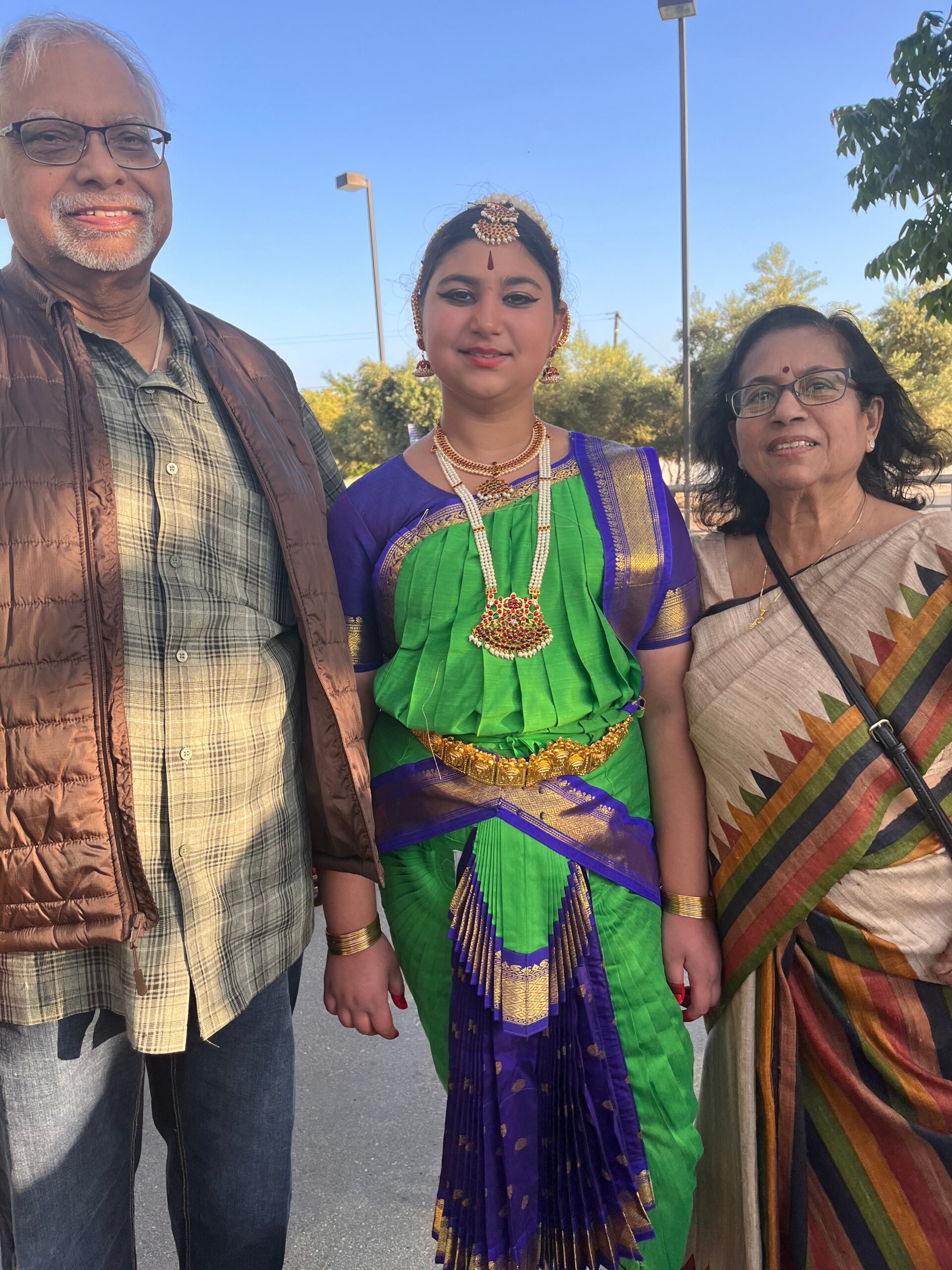 Three people posing outdoors: an older man in a brown vest, a young woman in green and purple traditional Indian dress with jewelry, and an older woman in a patterned sari smiling nearby.