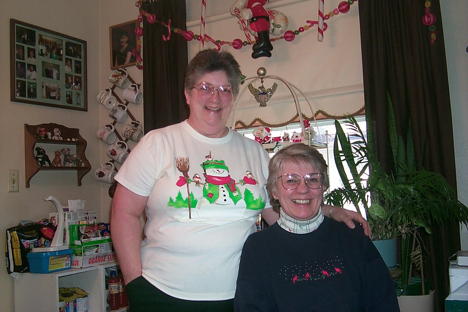 Two smiling women pose together indoors, one in a white Christmas-themed shirt and the other in a dark sweater, with holiday decorations in the background.
