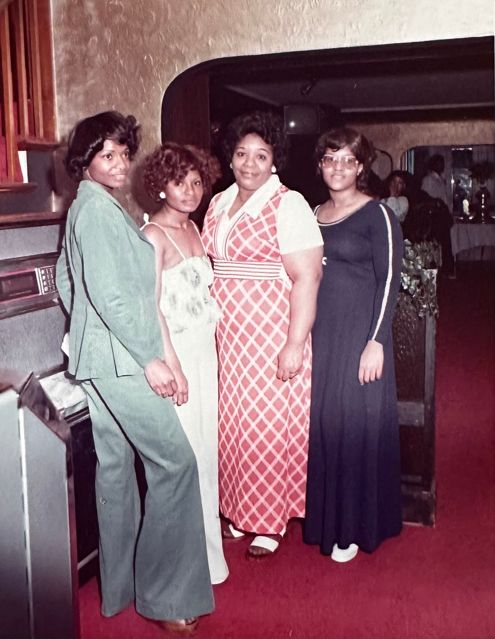 Four women posing together at a party in vintage 1970s outfits in a bar or club setting, smiling at the camera.