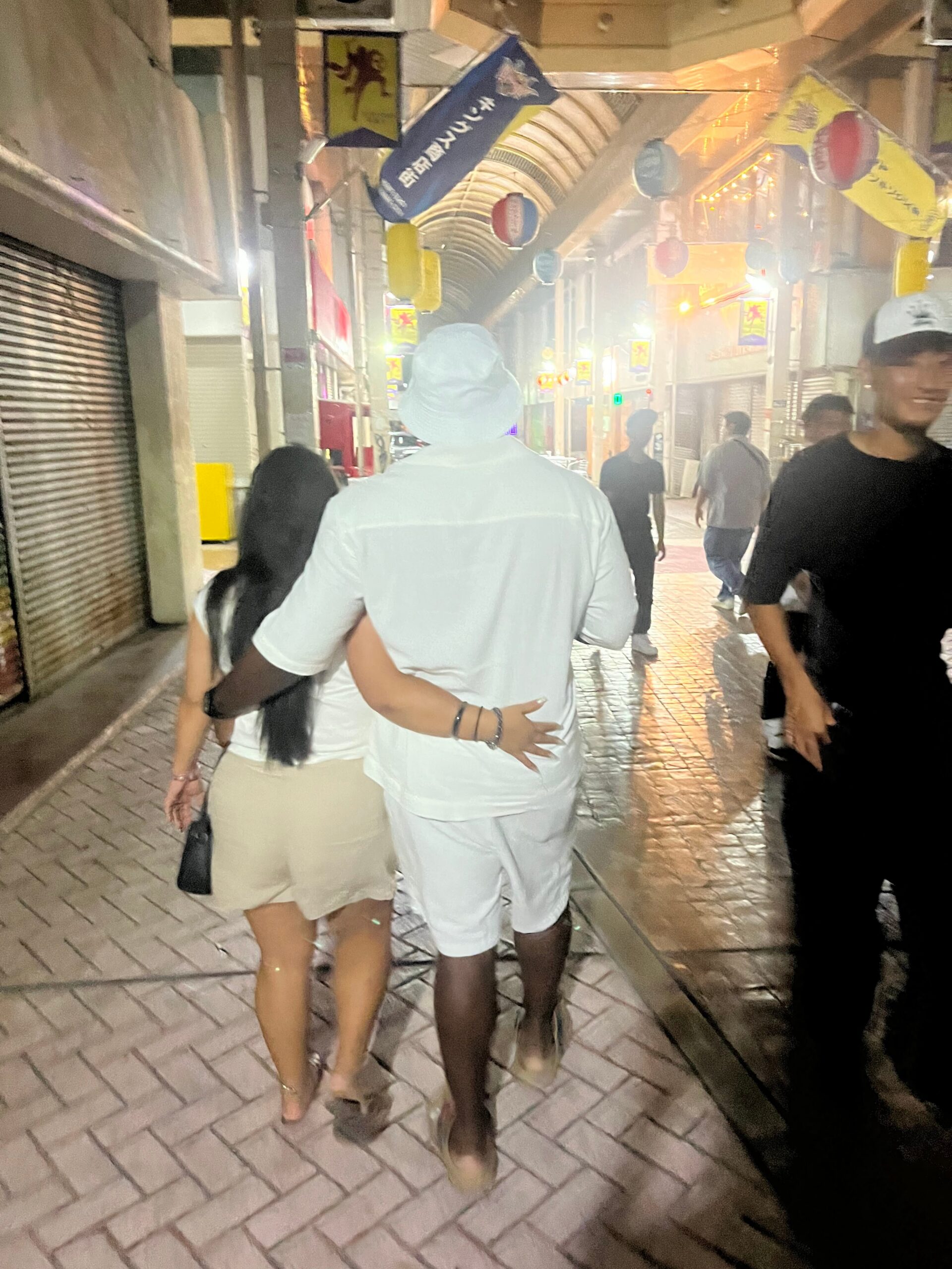 Back view of a couple walking arm in arm down a wet, lantern-lit alley at night.