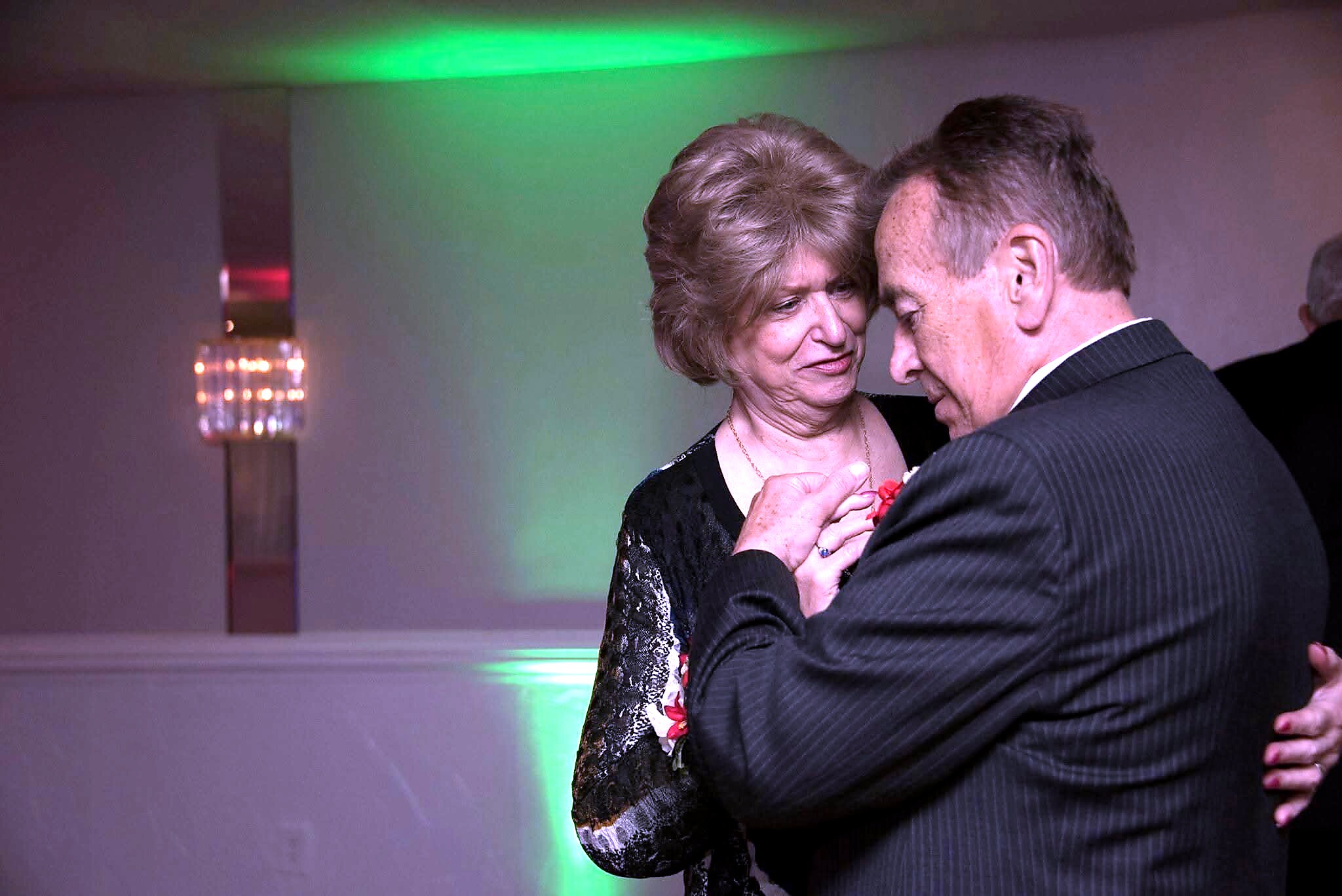 Couple dancing closely at a formal event, the man in a pinstriped suit embracing a woman in a black dress with floral details, under colorful party lighting.